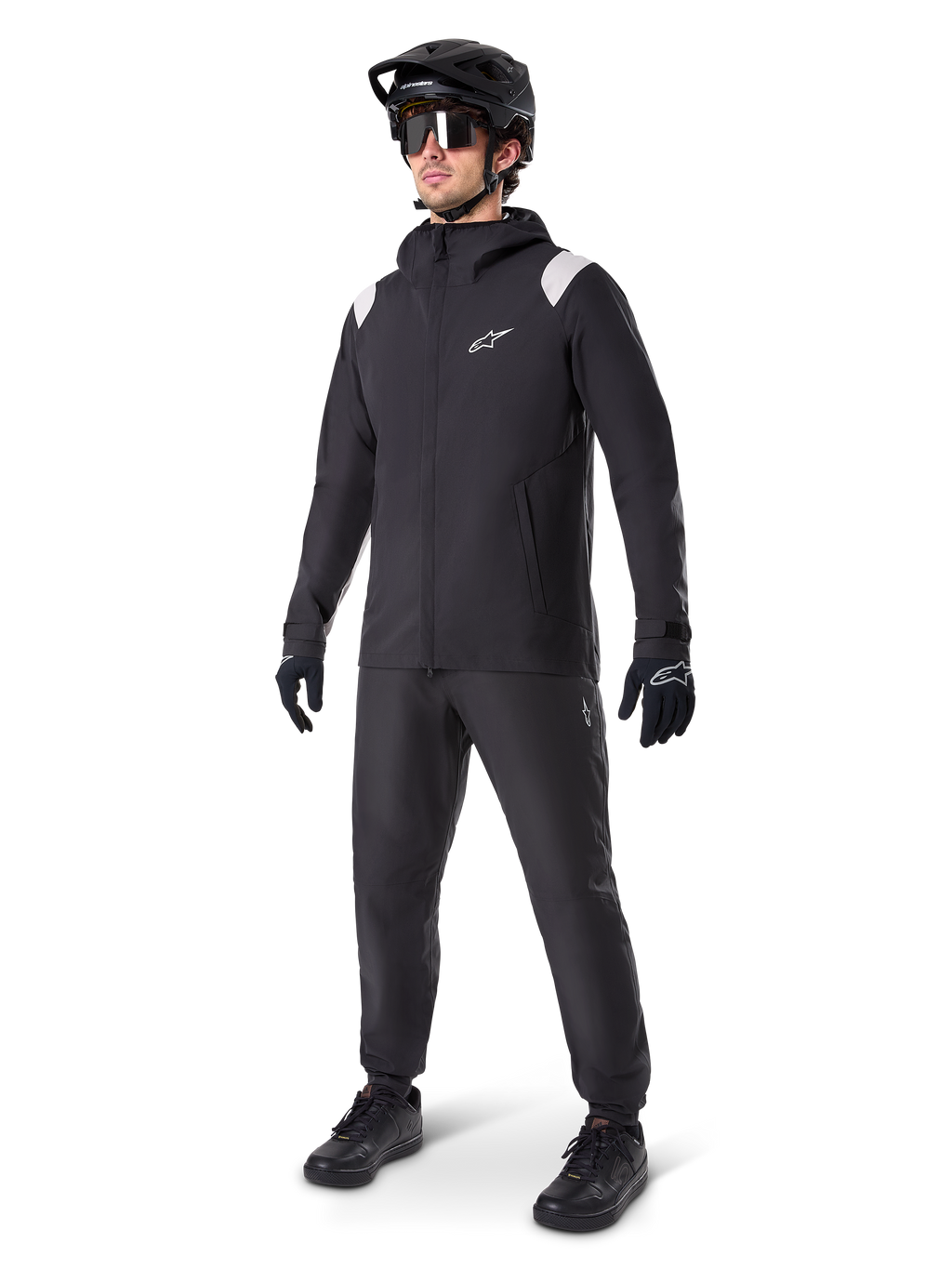 Alpinestars A-Dura Regenjacke, schwarze Mountainbike-Regenjacke mit weißen Akzenten an den Schultern, getragen von einem Fahrer mit passendem schwarzem MTB-Helm, Sonnenbrille, Handschuhen und Hose, technisches Allwetter-Radsport-Outfit