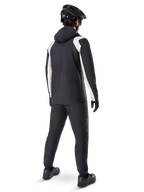 Alpinestars A-Dura Regenjacke, Rückansicht eines männlichen Radfahrers in der schwarz-weißen wasserdichten MTB-Jacke, dazu passende schwarze Radhose, schwarzer Helm und Handschuhe, technische Ausrüstung für das Mountainbiken bei nassen Bedingungen.