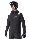 Alpinestars A-Dura Regenjacke, Mountainbike-Regenjacke, Schwarz mit hellgrauen Schulterakzenten, getragen von einem Fahrer mit schwarzem MTB-Helm und Sonnenbrille, weißes Logo auf der Brust, wasserdichte Konstruktion, kombiniert mit schwarzer Hose und Handschuhen für den Offroad-Radsport