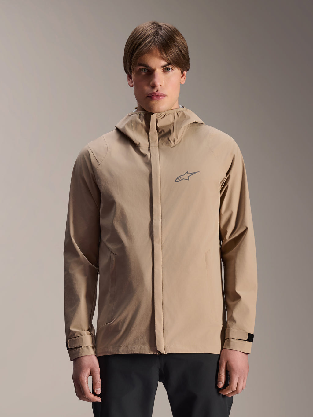 Alpinestars A-Dura Regenjacke, Hellbraune Mountainbike-Regenjacke mit Kapuze, von einem Model getragen, mit kleinem Brust-Logo, verstellbaren Ärmelbündchen und verdecktem Frontreißverschluss, wasserdichte Fahrradausrüstung