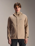 Alpinestars A-Dura Regenjacke, Hellbraune Mountainbike-Regenjacke mit Kapuze, von einem Model getragen, mit kleinem Brust-Logo, verstellbaren Ärmelbündchen und verdecktem Frontreißverschluss, wasserdichte Fahrradausrüstung