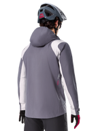 Alpinestars A-Dura Regenjacke, Mountainbike-Jacke, Dunkles Titangrau, Rückansicht mit hellgrauen Schulter- und Armeinsätzen, rotes Logo-Detail an der Seite, getragen mit einem grauen MTB-Helm und schwarz-roten Handschuhen, technische Trail-Bekleidung