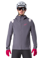 Alpinestars A-Dura Regenjacke, Mountainbike-Regenjacke, Dunkles Titanium-Grau, Kapuzendesign mit pinkem Alpinestars-Logo und hellgrauen Schultereinsätzen, getragen mit weißem MTB-Helm und leuchtend pinken Handschuhen, wasserdichte Ausrüstung für den Offroad-Radsport