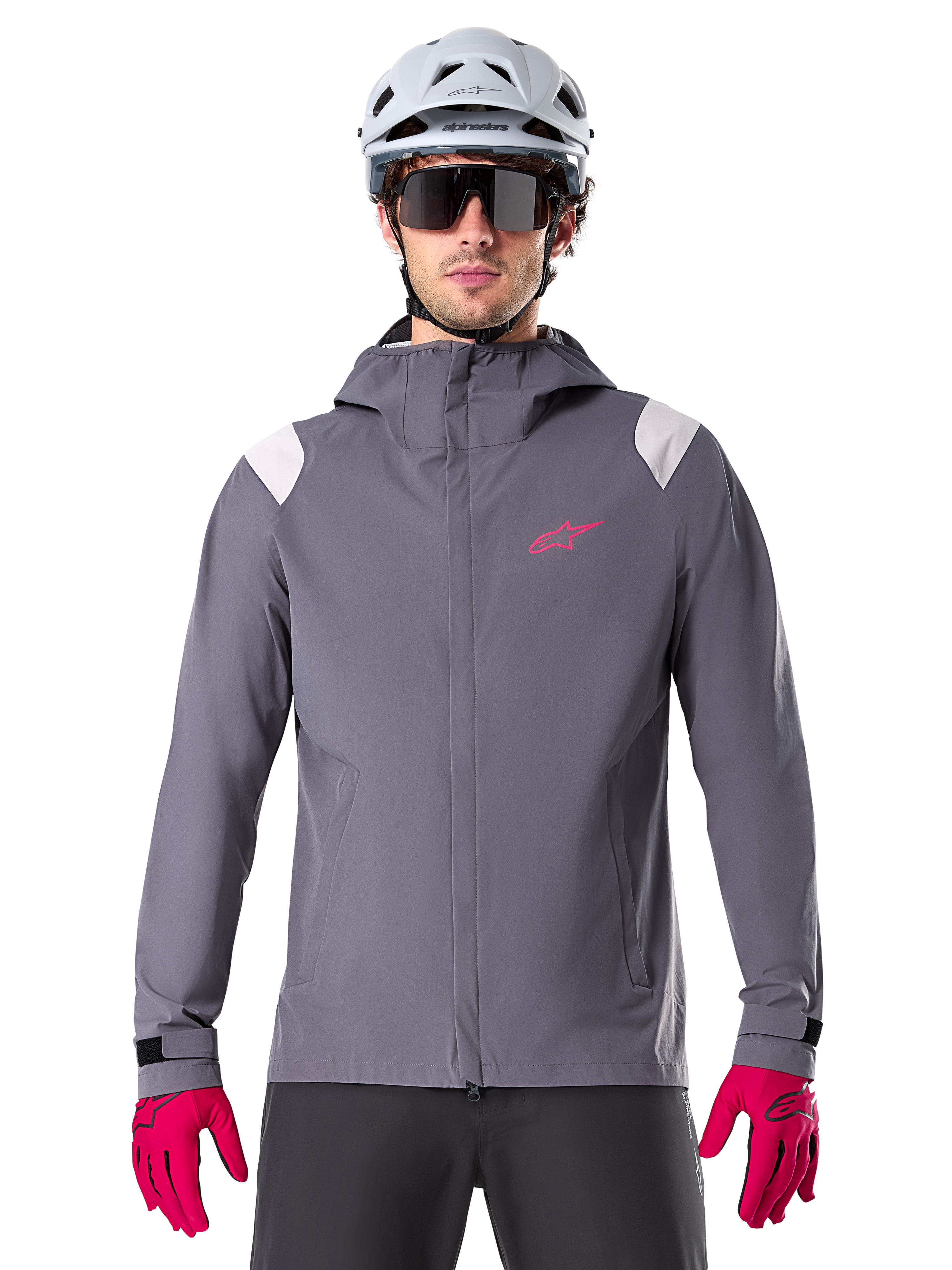 Alpinestars A-Dura Regenjacke, Mountainbike-Regenjacke, Dunkles Titanium-Grau, Kapuzendesign mit pinkem Alpinestars-Logo und hellgrauen Schultereinsätzen, getragen mit weißem MTB-Helm und leuchtend pinken Handschuhen, wasserdichte Ausrüstung für den Offroad-Radsport