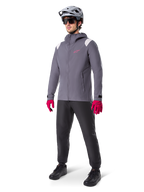 Männliches Model in der Alpinestars A-Dura Regenjacke in Dark Titanium, graue Kapuzenjacke mit pinkem Brustlogo und hellgrauen Schultereinsätzen, kombiniert mit einem grauen MTB-Helm, dunkler Sonnenbrille, leuchtend pinken Handschuhen und einer schwarzen Hose für den Offroad-Radsport.