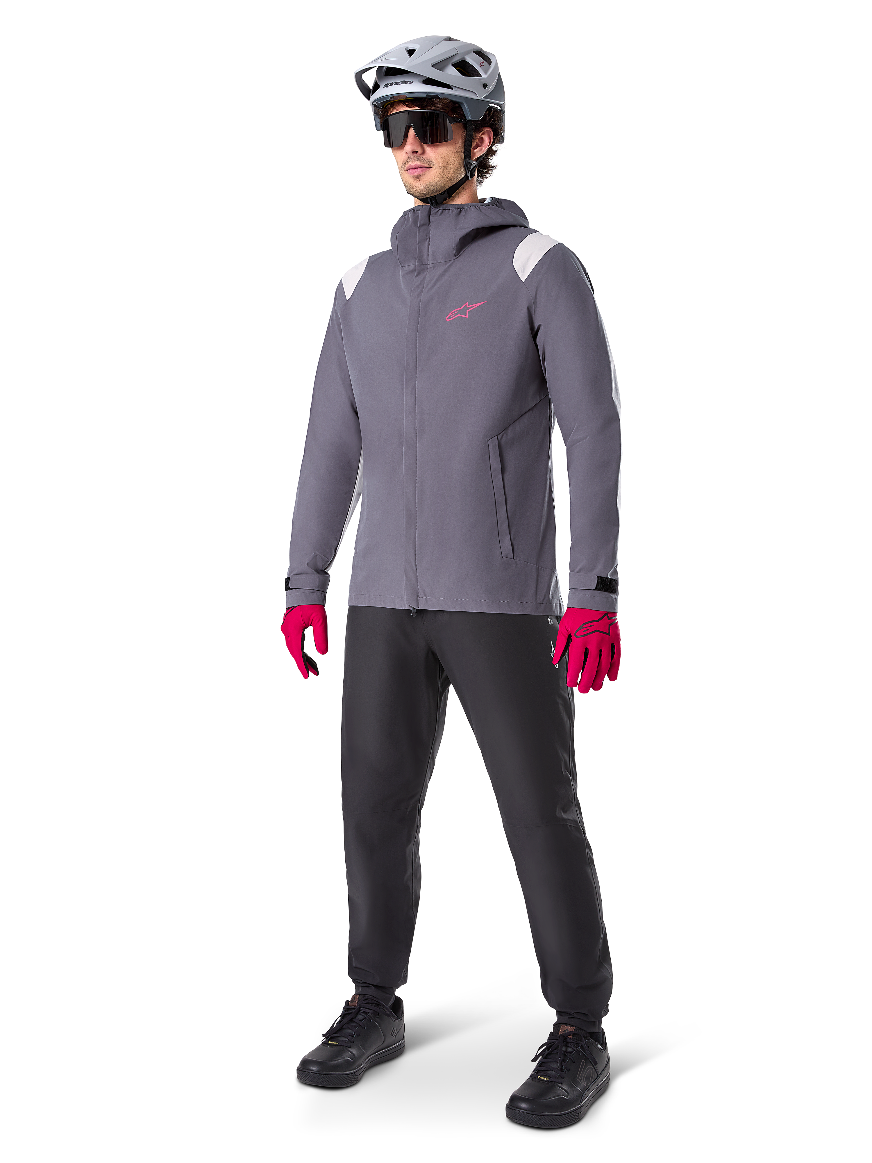 Männliches Model in der Alpinestars A-Dura Regenjacke in Dark Titanium, graue Kapuzenjacke mit pinkem Brustlogo und hellgrauen Schultereinsätzen, kombiniert mit einem grauen MTB-Helm, dunkler Sonnenbrille, leuchtend pinken Handschuhen und einer schwarzen Hose für den Offroad-Radsport.