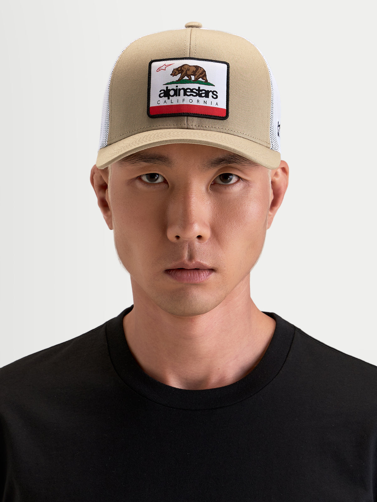 Alpinestars Cali 2.0 Kappe, Sportswear Trucker-Kappe in Khaki und Weiß, getragen von einem Model, mit gebogenem Schirm, strukturierter Twill-Front, atmungsaktiver Mesh-Rückseite und California-Bären-Patch auf der Vorderseite