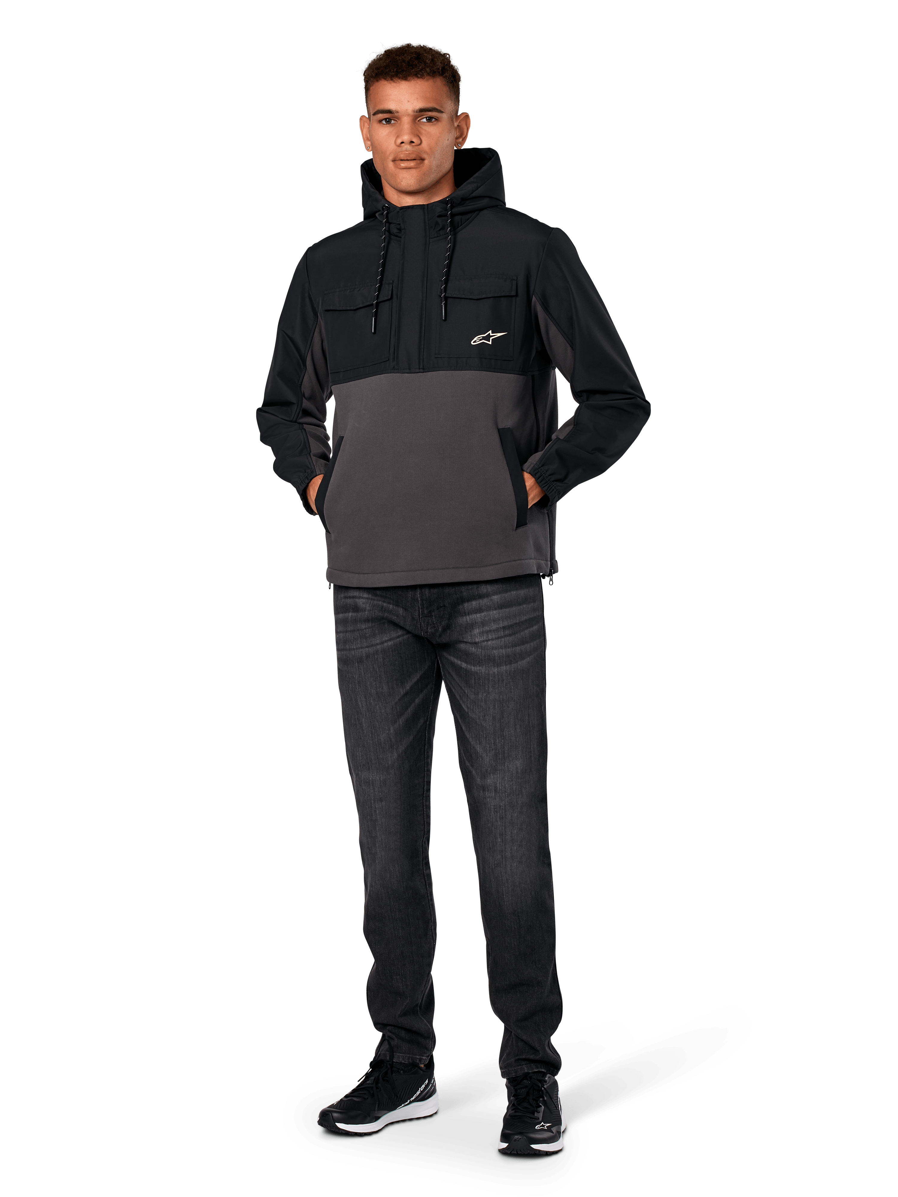 Alpinestars Juncture Hybrid Jacke, Schwarz und Gunmetal, Anorak-Kapuzenpullover aus Mischmaterial mit Canvas-Fleece und Ripstop-Overlays, weißes A-Star Logo auf der Brust, Kapuze mit Kordelzug, Modell trägt dazu dunkle Jeans und schwarze Sneaker, lässige vom Motorsport inspirierte Streetwear