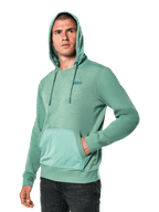 Alpinestars Rooted Hoodie, grünes Slub-French-Terry-Sweatshirt mit Überzugskapuze und Kängurutasche, kleines STARS-Logo auf der Brust, getragen von einem Model mit aufgesetzter Kapuze und Hand in der Tasche, lässige, vom Motorsport inspirierte Streetwear