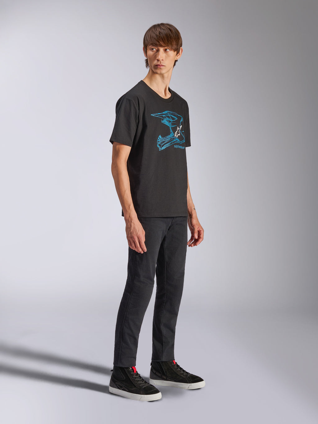 Alpinestars CSF Tee, schwarzes Kurzarm-T-Shirt mit blauer MX-Helm-Grafik, getragen von einem Model mit schwarzer Hose und High-Top-Sneakern, lässige Motorsport-Streetwear