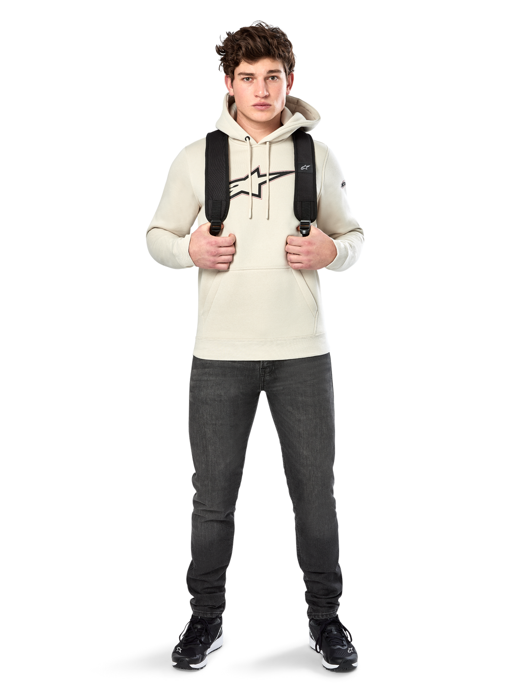 Alpinestars Insignia Hoodie, legere Streetwear, Fleece-Pullover in Off-White, Logo in Carbonfaser-Optik mit roter Umrandung, Kapuze mit Kordelzug, Kängurutasche, getragen von einem Model mit schwarzem Rucksack und dunkelgrauen Jeans