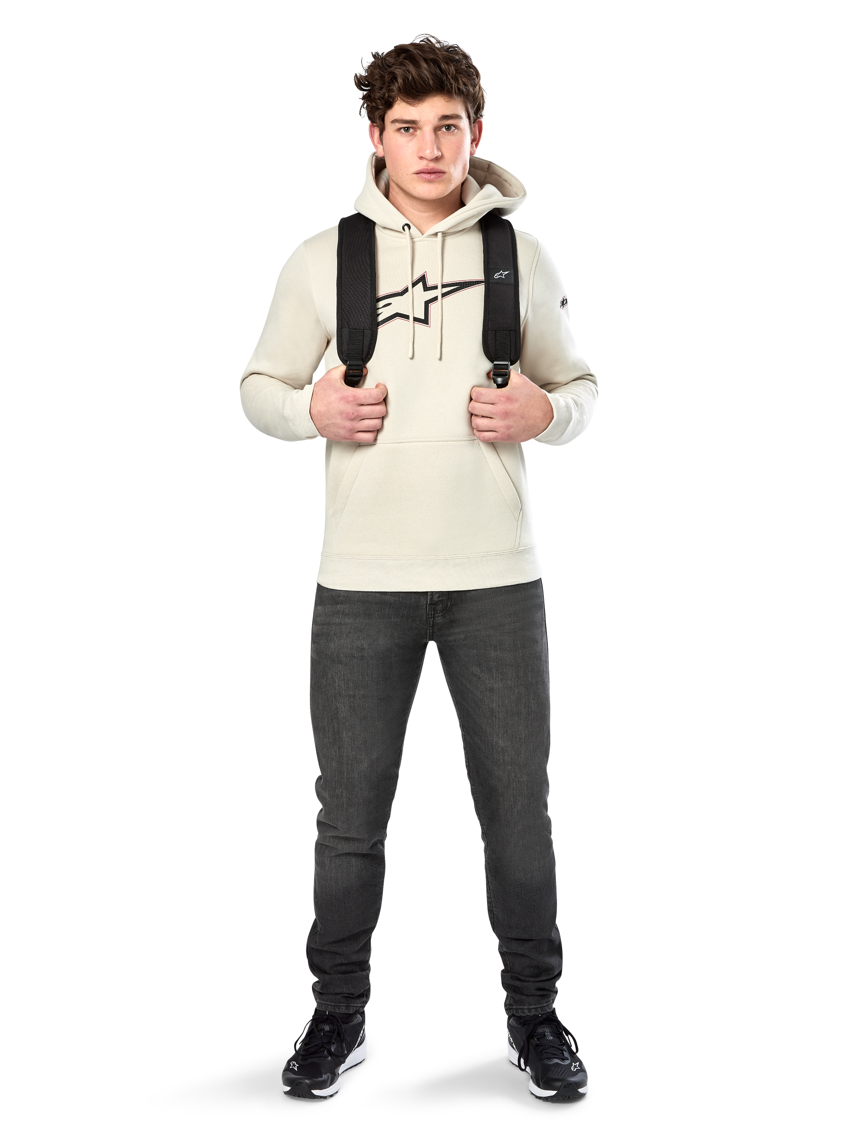 Alpinestars Insignia Hoodie, legere Streetwear, Fleece-Pullover in Off-White, Logo in Carbonfaser-Optik mit roter Umrandung, Kapuze mit Kordelzug, Kängurutasche, getragen von einem Model mit schwarzem Rucksack und dunkelgrauen Jeans