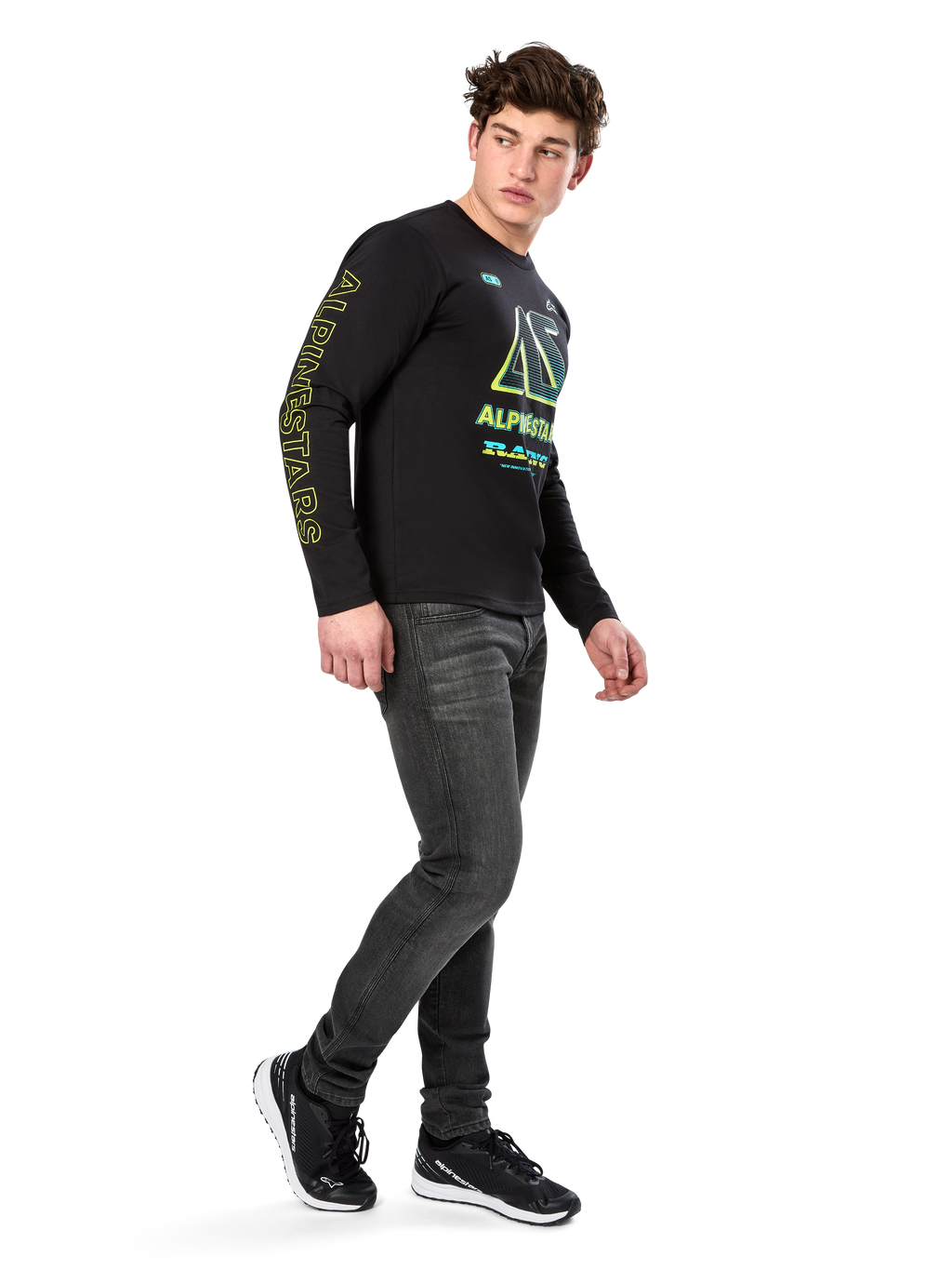 Mann trägt Alpinestars Ayess CSF Langarm-T-Shirt, Schwarz, mit gelber Ärmelbeschriftung und leuchtenden limetten- und cyanfarbenen Brustgrafiken, kombiniert mit dunkelgrauen Jeans und schwarzen Sneakern, lässige Motorsport-inspirierte Streetwear