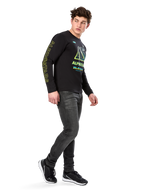 Mann trägt Alpinestars Ayess CSF Langarm-T-Shirt, Schwarz, mit gelber Ärmelbeschriftung und leuchtenden limetten- und cyanfarbenen Brustgrafiken, kombiniert mit dunkelgrauen Jeans und schwarzen Sneakern, lässige Motorsport-inspirierte Streetwear