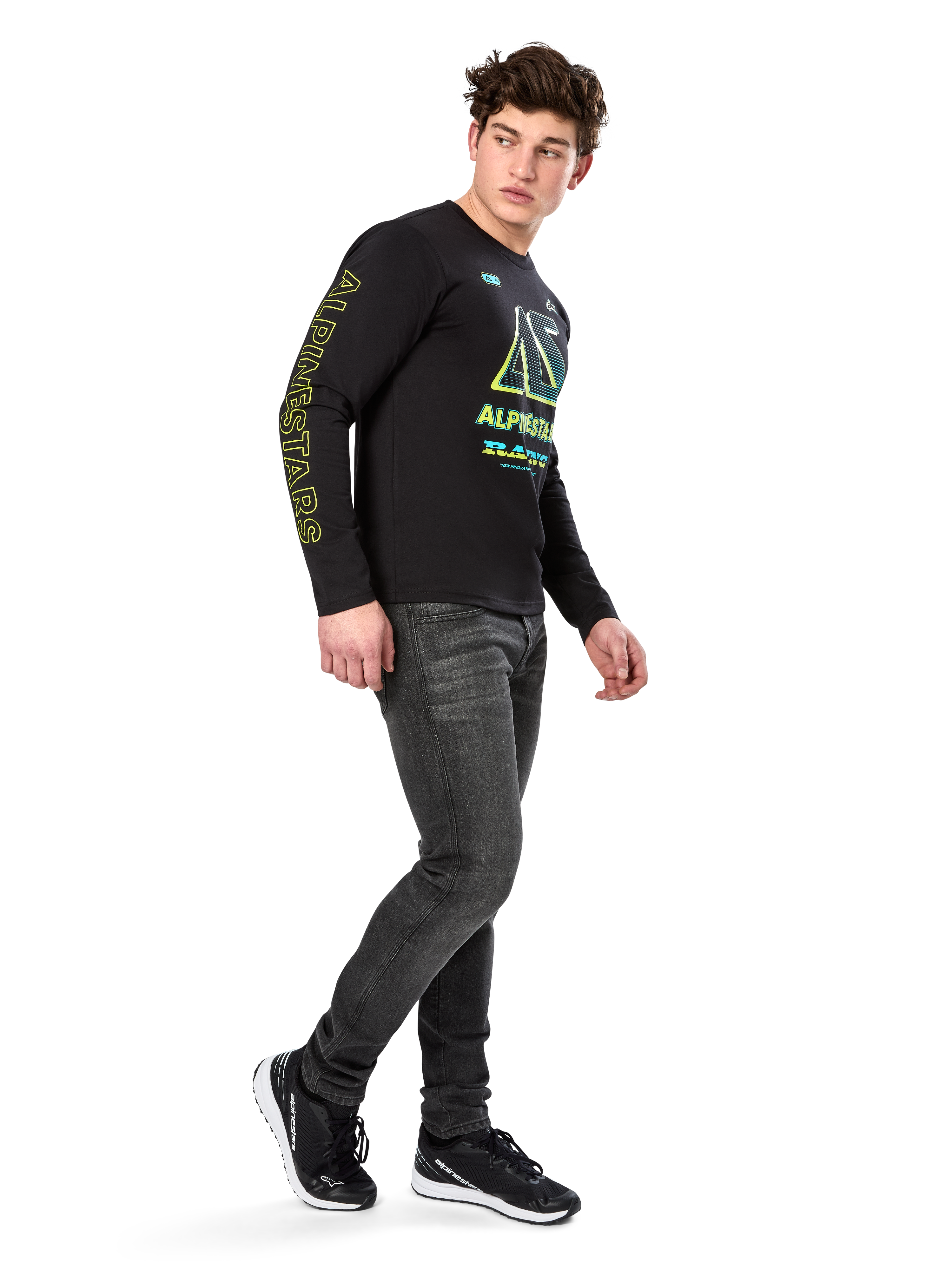 Mann trägt Alpinestars Ayess CSF Langarm-T-Shirt, Schwarz, mit gelber Ärmelbeschriftung und leuchtenden limetten- und cyanfarbenen Brustgrafiken, kombiniert mit dunkelgrauen Jeans und schwarzen Sneakern, lässige Motorsport-inspirierte Streetwear
