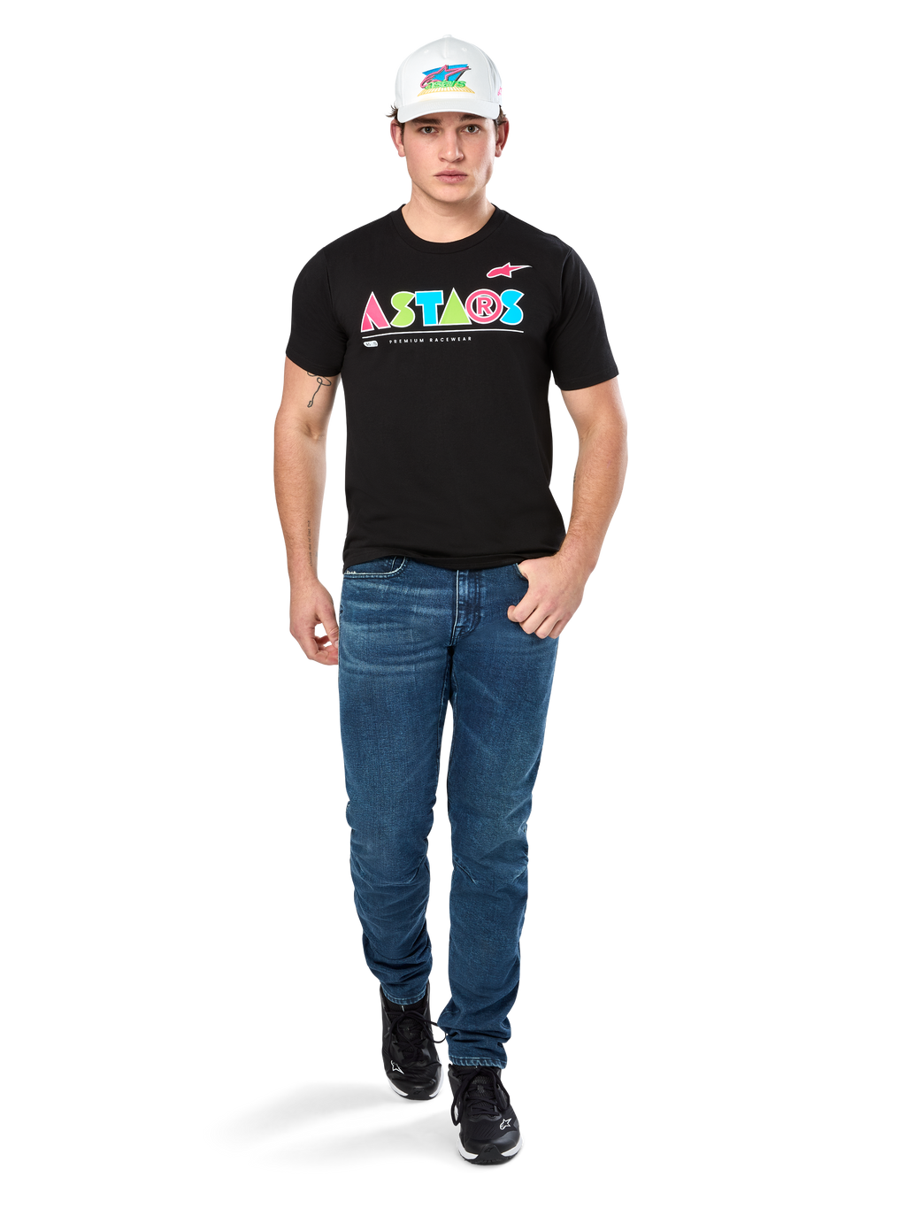 Männliches Model im Alpinestars Cereal CSF Tee, schwarzes Kurzarm-T-Shirt mit farbenfrohem geometrischem ASTARS-Logo, kombiniert mit blauen Jeans und weißer Kappe, lässige motorsport-inspirierte Streetwear.