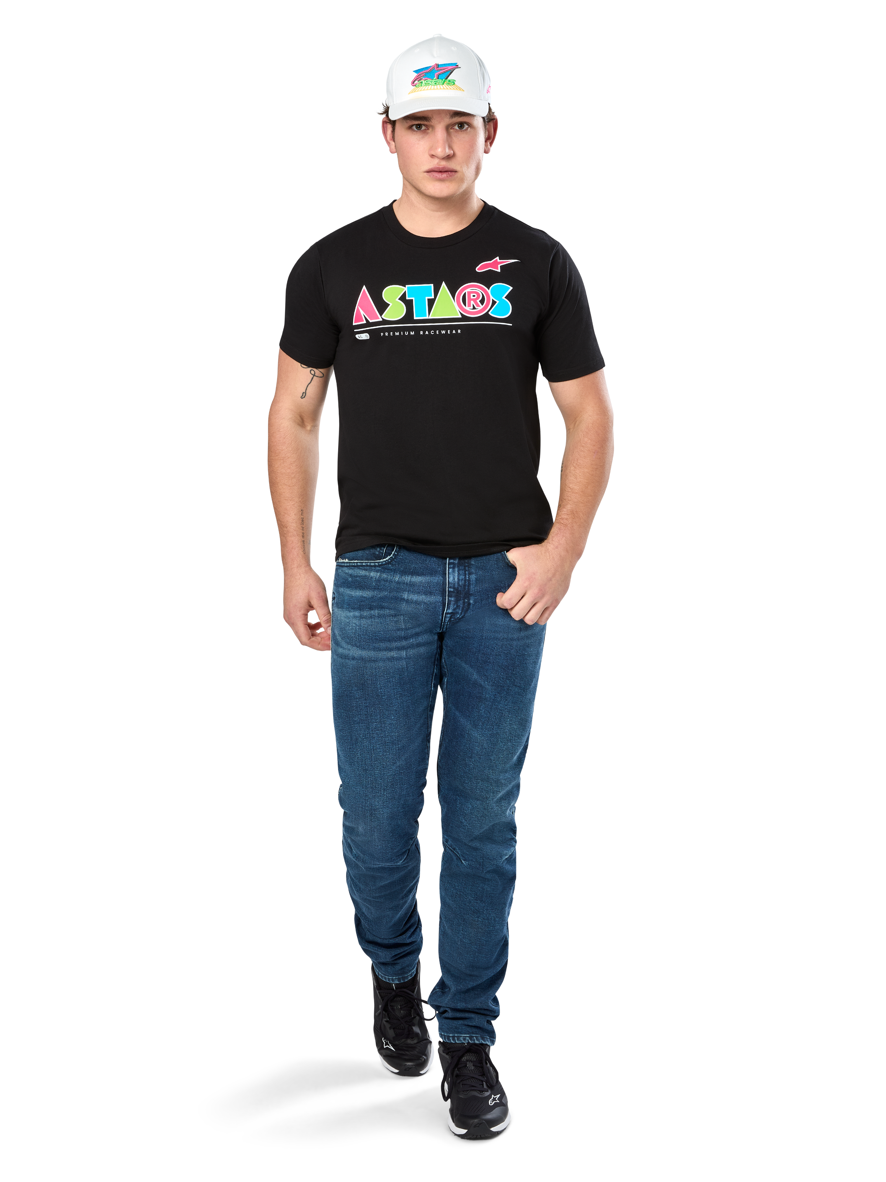 Männliches Model im Alpinestars Cereal CSF Tee, schwarzes Kurzarm-T-Shirt mit farbenfrohem geometrischem ASTARS-Logo, kombiniert mit blauen Jeans und weißer Kappe, lässige motorsport-inspirierte Streetwear.