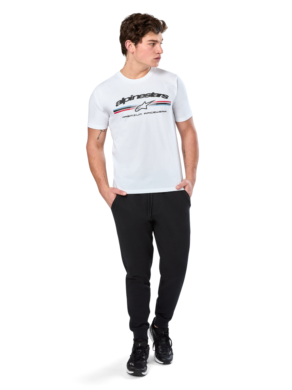 Alpinestars Prevail CSF Tee, Kurzarm-T-Shirt, Weiß, mit dreifarbigem Streifenlogo im Paddock-Stil und Alpinestars-Branding auf der Vorderseite, getragen von einem Model mit schwarzen Jogginghosen und schwarzen Sneakern, lässige Motorsport-Streetwear