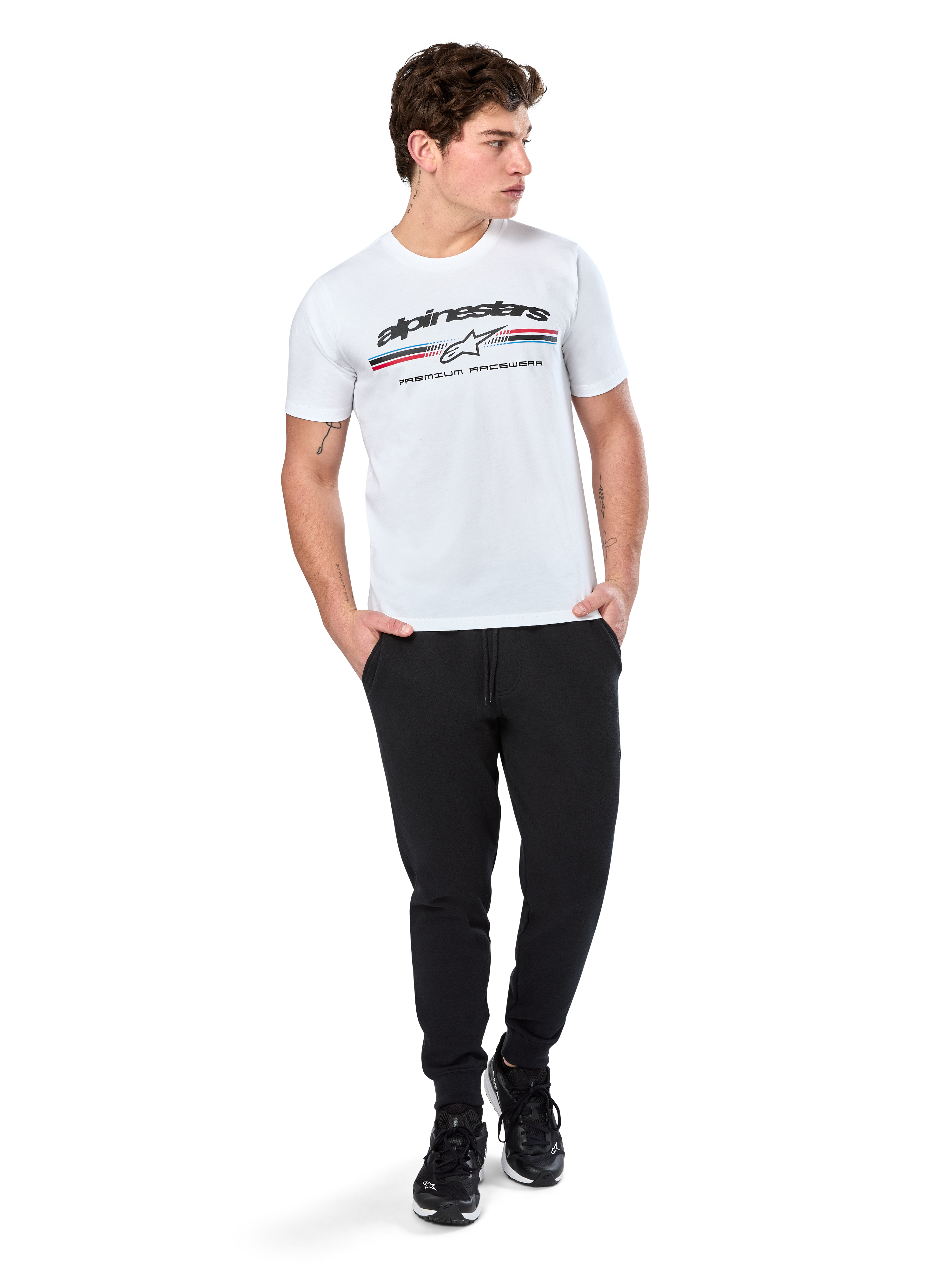 Alpinestars Prevail CSF Tee, Kurzarm-T-Shirt, Weiß, mit dreifarbigem Streifenlogo im Paddock-Stil und Alpinestars-Branding auf der Vorderseite, getragen von einem Model mit schwarzen Jogginghosen und schwarzen Sneakern, lässige Motorsport-Streetwear