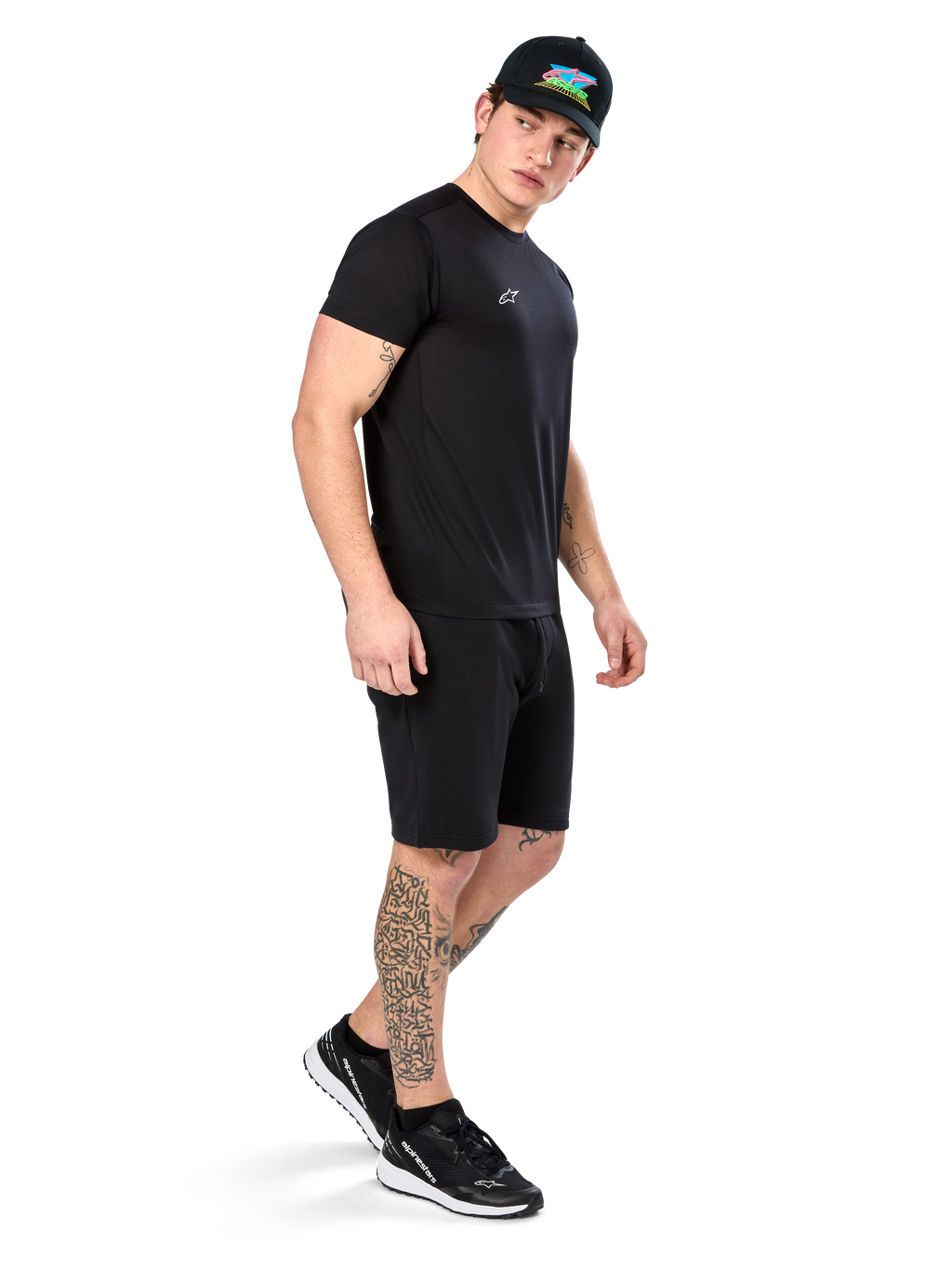 Mann mit Tattoos im Alpinestars Point Performance Tee, Kurzärmliges schwarzes Performance-T-Shirt, kleines weißes Logo, kombiniert mit schwarzen Shorts, einer schwarzen Kappe mit bunter Grafik und schwarz-weißen Sneakern, sportlich-lässiger Lifestyle