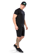 Mann mit Tattoos im Alpinestars Point Performance Tee, Kurzärmliges schwarzes Performance-T-Shirt, kleines weißes Logo, kombiniert mit schwarzen Shorts, einer schwarzen Kappe mit bunter Grafik und schwarz-weißen Sneakern, sportlich-lässiger Lifestyle