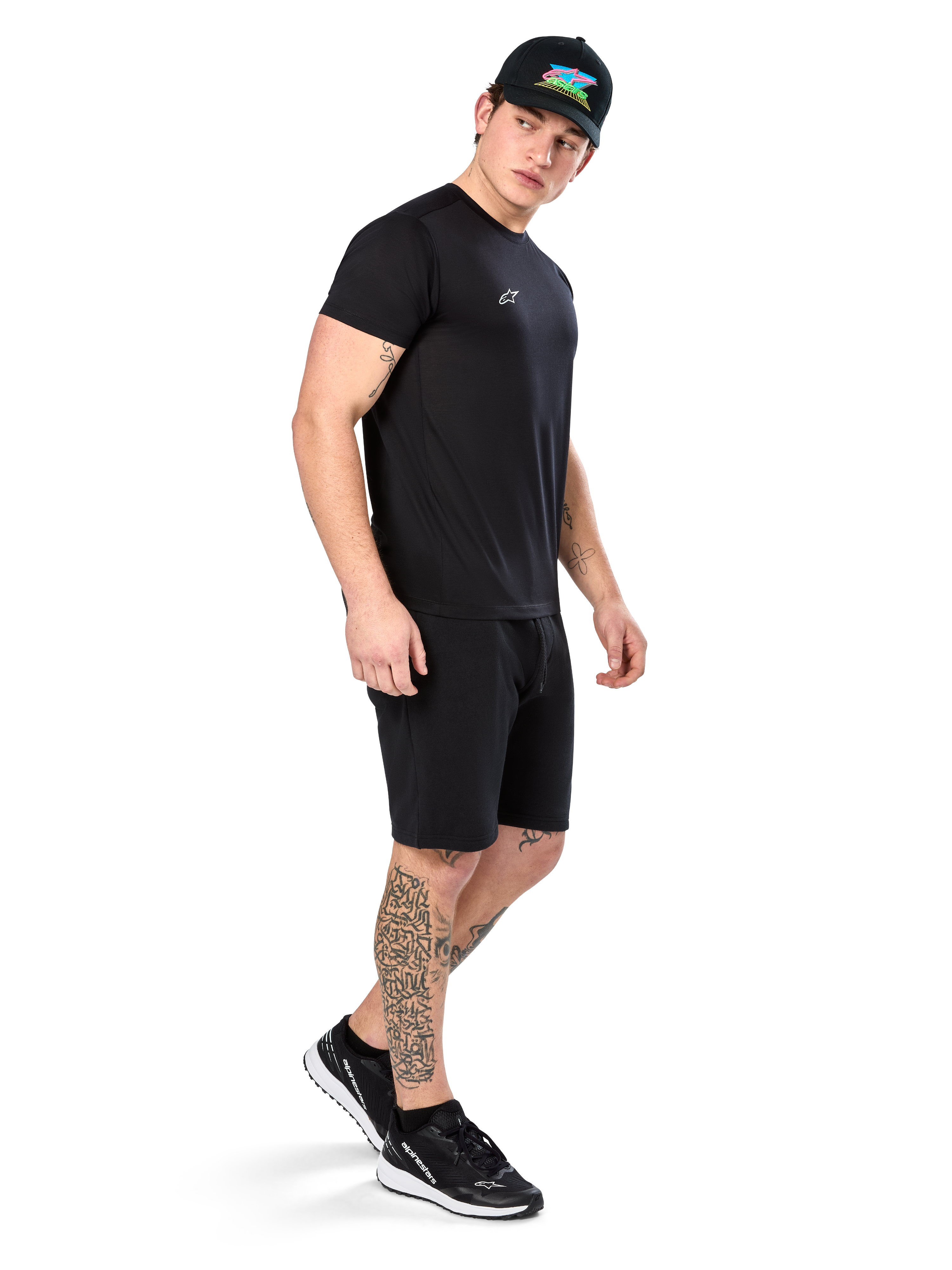Mann mit Tattoos im Alpinestars Point Performance Tee, Kurzärmliges schwarzes Performance-T-Shirt, kleines weißes Logo, kombiniert mit schwarzen Shorts, einer schwarzen Kappe mit bunter Grafik und schwarz-weißen Sneakern, sportlich-lässiger Lifestyle