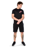 Model trägt Alpinestars Attrition Performance Tee, kurzärmliges Performance-T-Shirt, Schwarz mit pinkem Astars-Logo auf der Brust, kombiniert mit schwarzen Shorts und Sneakern, lässige motorsport-inspirierte Sportbekleidung