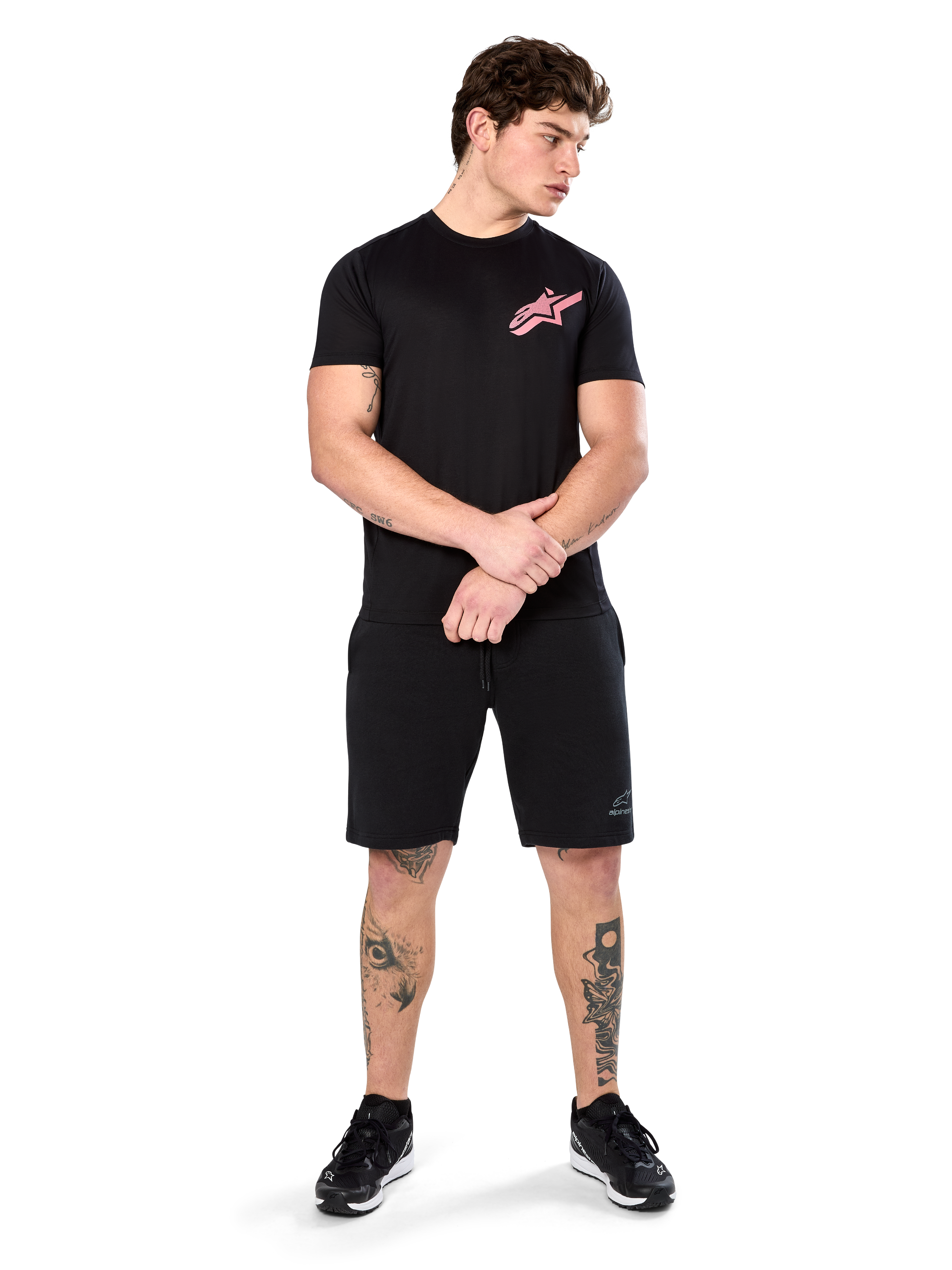Model trägt Alpinestars Attrition Performance Tee, kurzärmliges Performance-T-Shirt, Schwarz mit pinkem Astars-Logo auf der Brust, kombiniert mit schwarzen Shorts und Sneakern, lässige motorsport-inspirierte Sportbekleidung