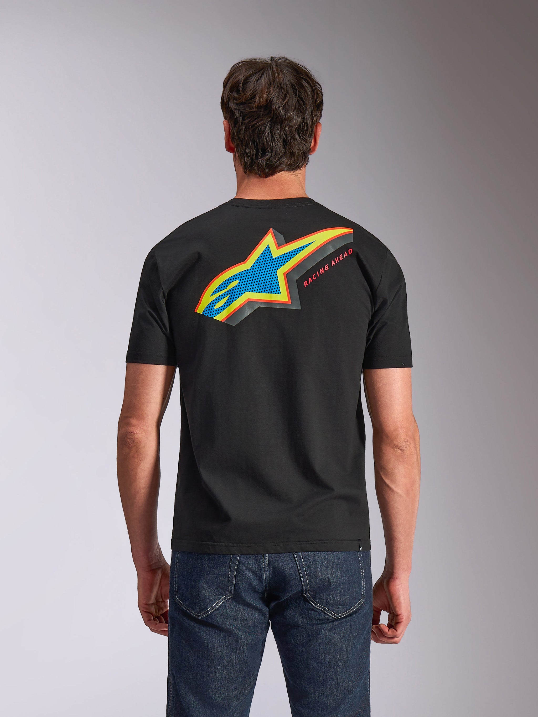 Alpinestars Angler CSF Tee, Short Sleeve T-Shirt, Black, back view showing vibrant multi-color Astars logo with perforated fill and 'RACING AHEAD' text, worn with blue jeans, casual motorsport-inspired apparel