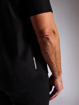 Alpinestars Strive Woven Shirt, schwarzes kurzärmeliges Hemd mit Knopfleiste, vertikales weißes Alpinestars-Logo an der Seite, Detailaufnahme vom unteren Saum und Markenlogo, lässige Motorsport-Streetwear