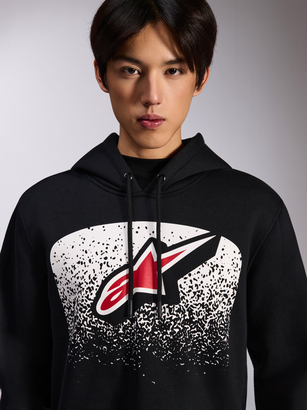 Alpinestars Obsessed Hoodie, schwarzer Fleece-Kapuzenpullover, mit einem rot-weißen Astars-Logo in einer weiß gepunkteten MX-Verlaufsgrafik auf der Brust, von einem Model getragen, lässige Motorsport-Streetwear mit verstellbarem Kordelzug an der Kapuze