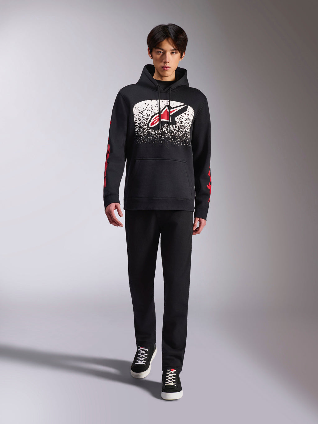 Alpinestars Obsessed Hoodie, schwarzer Kapuzenpullover, großes weißes und rotes Astars-Logo mit MX-Farbverlauf, rote Wortmarken an den Ärmeln, getragen von einem Model in einem komplett schwarzen Freizeitoutfit, motorsport-inspirierte Lifestyle-Bekleidung