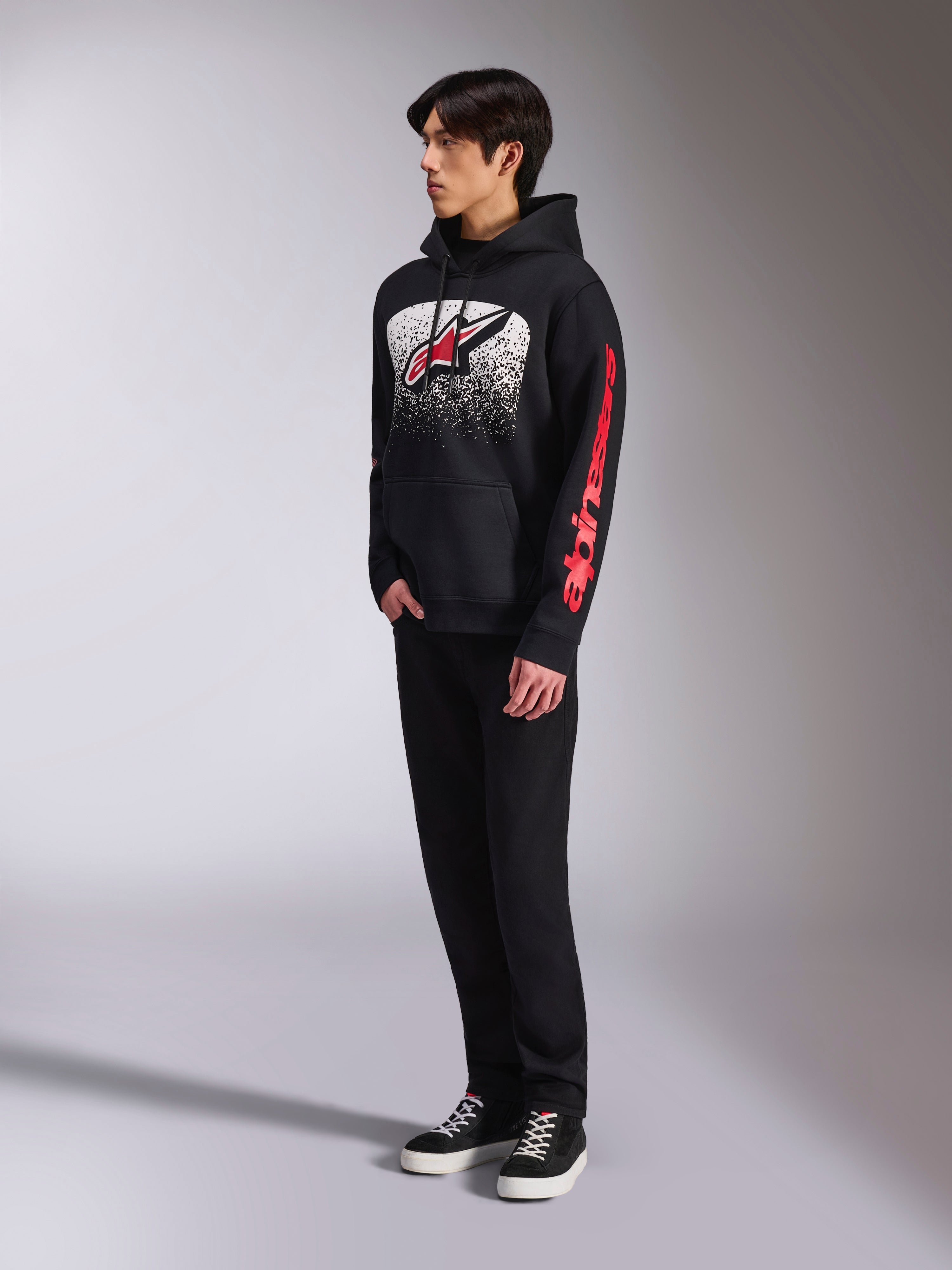 Alpinestars Obsessed Hoodie, schwarzer Kapuzenpullover aus Fleece, mit einer großen weiß gesprenkelten Grafik auf der Brust, rotem Astars-Logo und rotem Branding an den Ärmeln, getragen von einem Model mit schwarzer Hose und Sneakern, lässige Motorsport-Streetwear