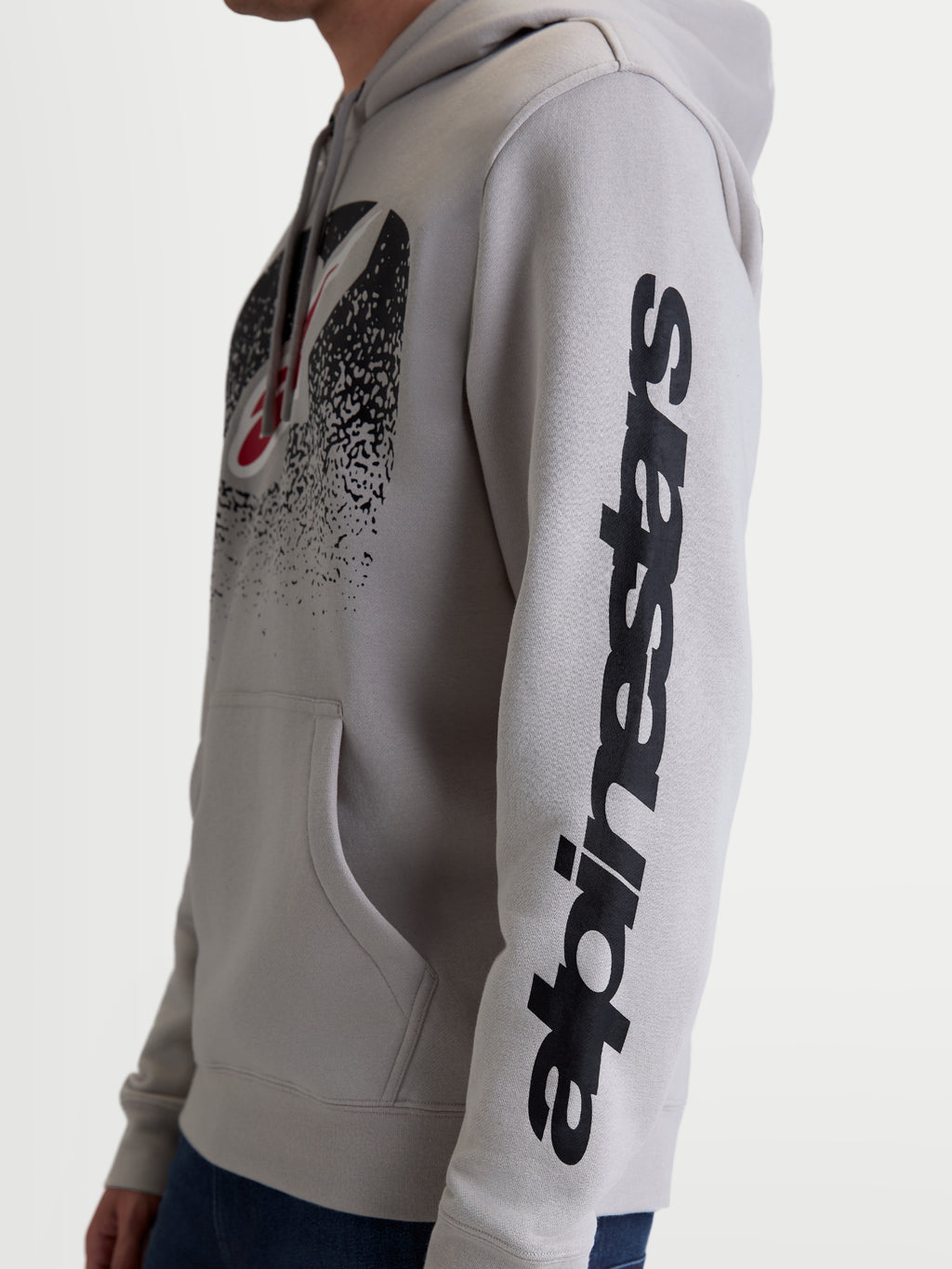 Alpinestars Obsessed Hoodie, lässiger Motorsport-Kapuzenpullover, Unisex, Zementgrau und Schwarz, Seitenansicht am Model mit Fokus auf das vertikale Alpinestars-Ärmellogo und die MX-inspirierte Brustgrafik mit Farbverlauf, aus Baumwoll-Fleece und recyceltem Polyester.