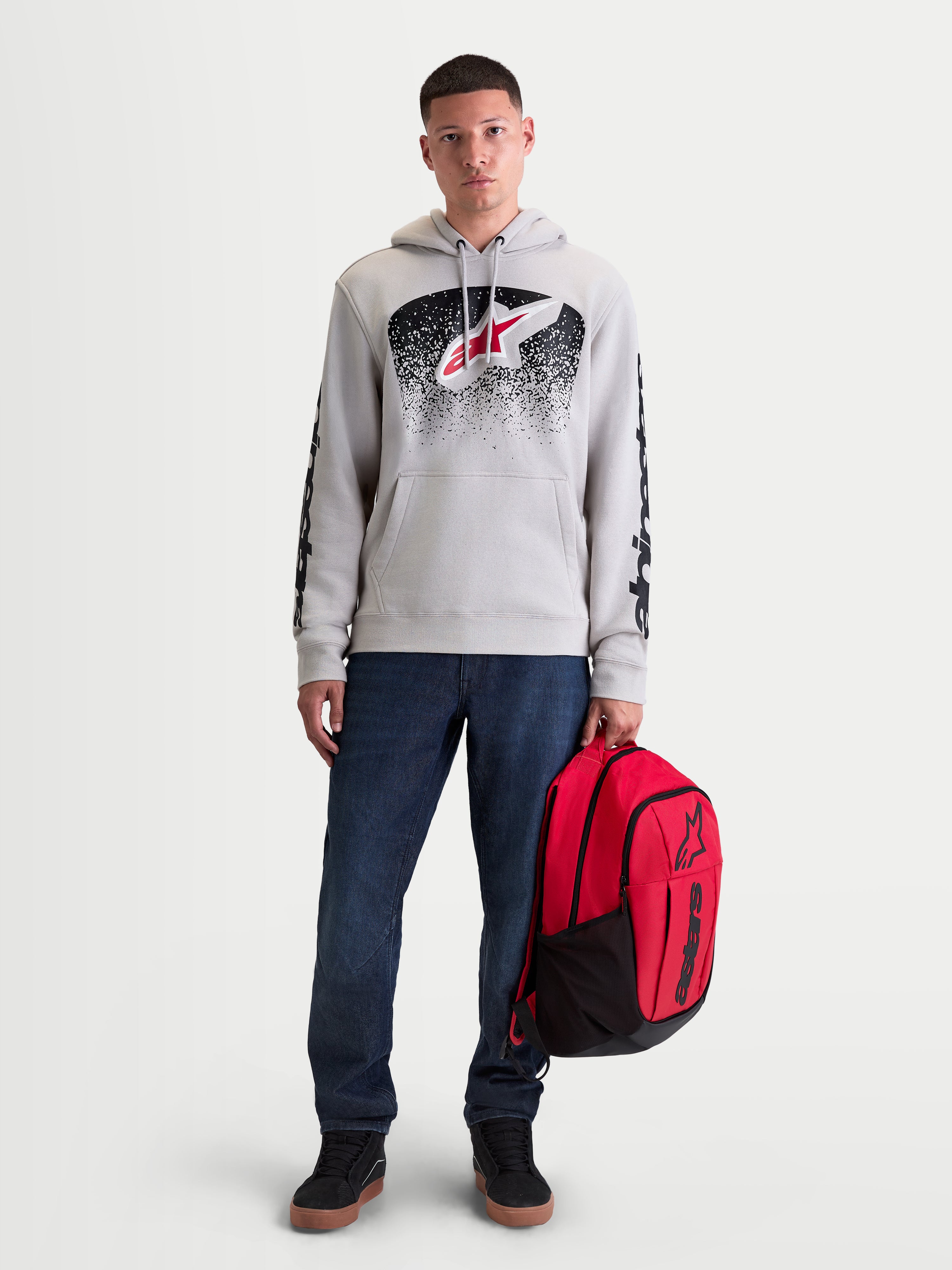 Alpinestars Obsessed Hoodie, Sportswear-Kapuzenpullover für Herren, Zementgrau, Rot und Schwarz, MX-inspirierte Verlaufsgrafiken und Ärmellogos. Gezeigt an einem männlichen Modell, das einen roten Alpinestars Rucksack hält.