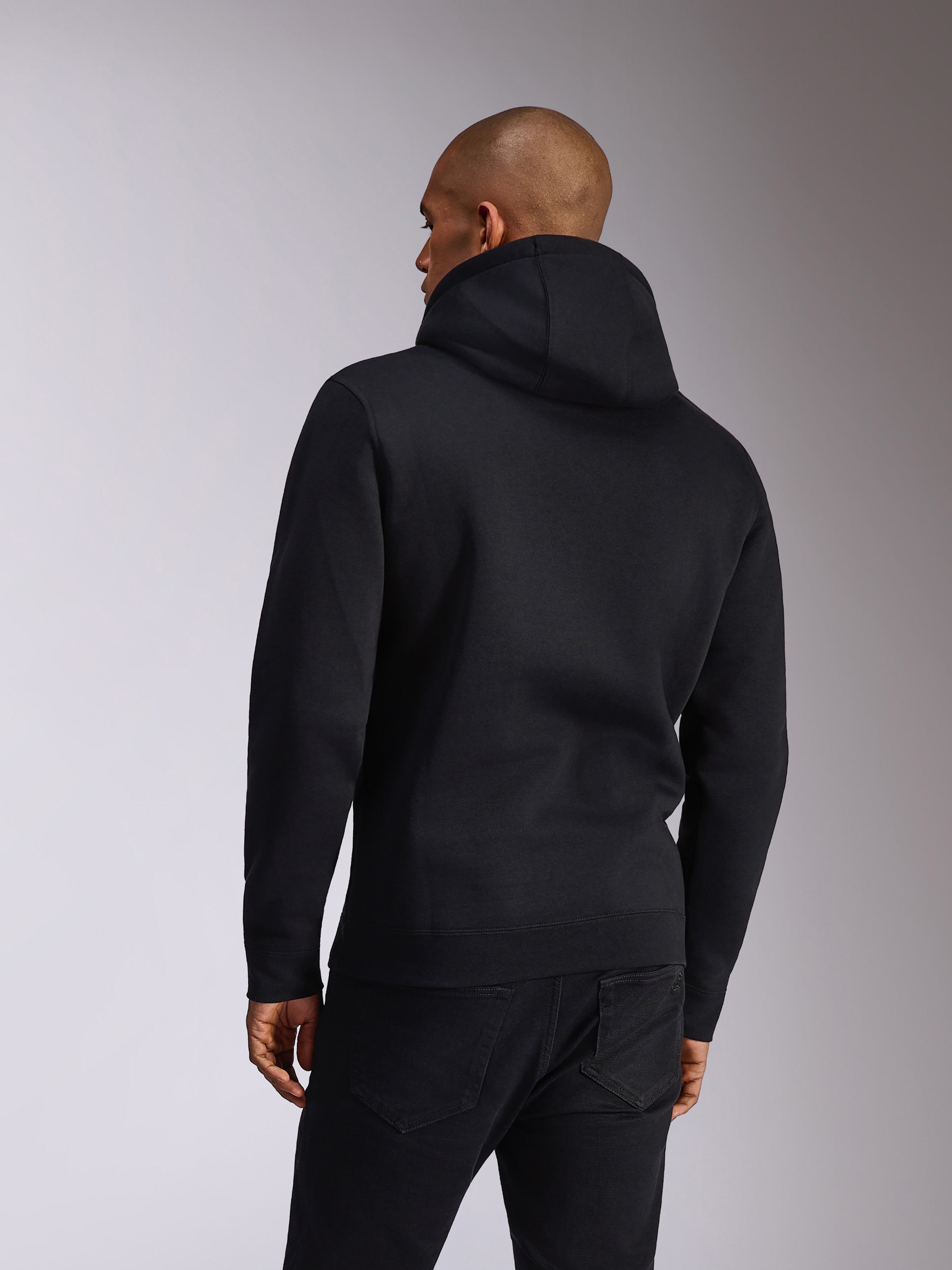 Alpinestars Ellipsoid Hoodie, Schwarz, Rückansicht einer Person, die einen Fleece-Kapuzenpullover trägt, lange Ärmel mit Rippbündchen an Ärmeln und Saum, kombiniert mit schwarzen Jeans, lässige Motorsport-inspirierte Sportbekleidung