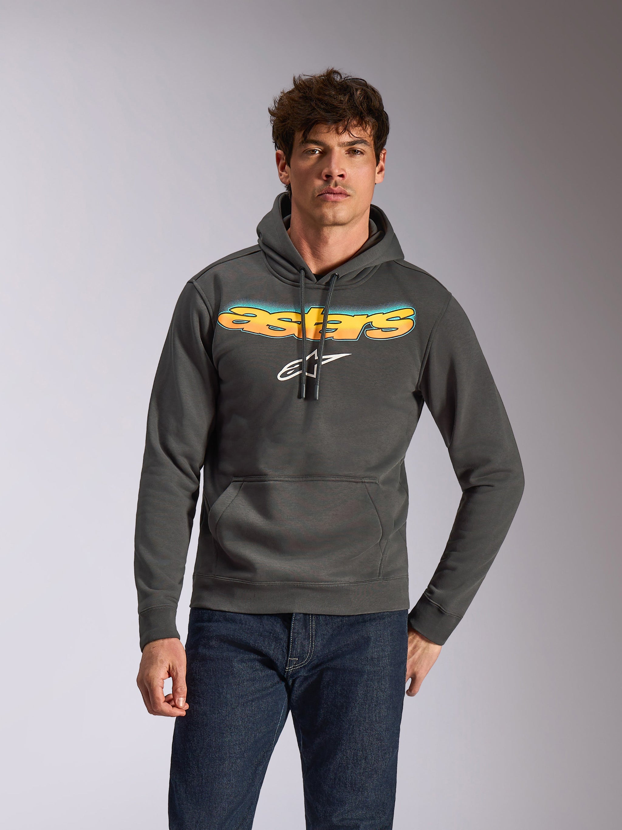 Alpinestars Ellipsoid Hoodie, lässige Sportbekleidung, Gunmetal-Grau mit gelbem und blauem Astars-Schriftzug mit Farbverlauf und weißem Logo, Kapuzenpullover mit Kordelzug, getragen mit dunkelblauen Jeans