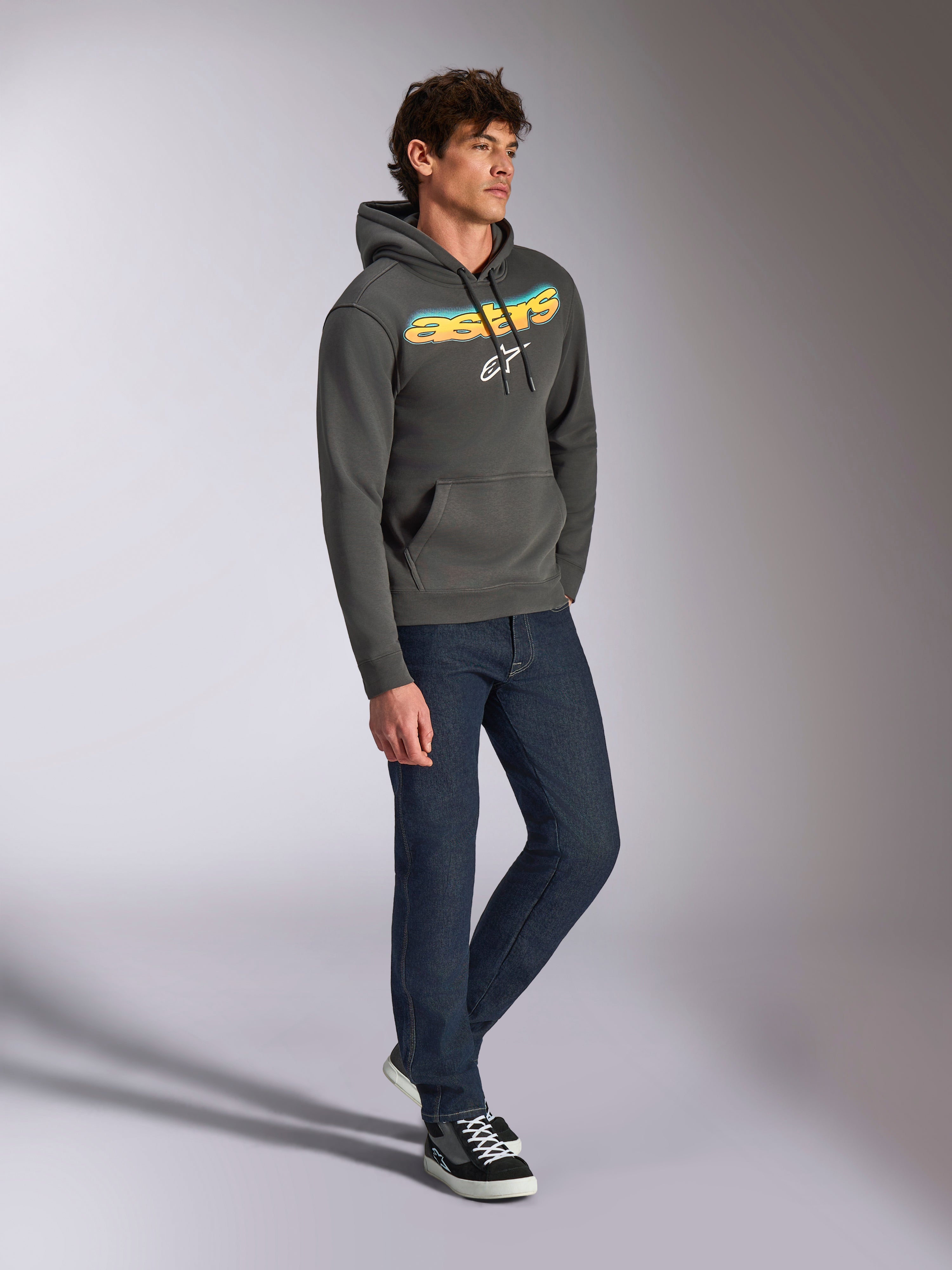 Alpinestars Ellipsoid Hoodie, Fleece-Kapuzenpullover in Gunmetal-Grau, vorne gebogener Astars-Schriftzug mit blau-gelbem Farbverlauf und weißem Logo, kombiniert mit dunkler Jeans und schwarzen Sneakern, lässige Streetwear.