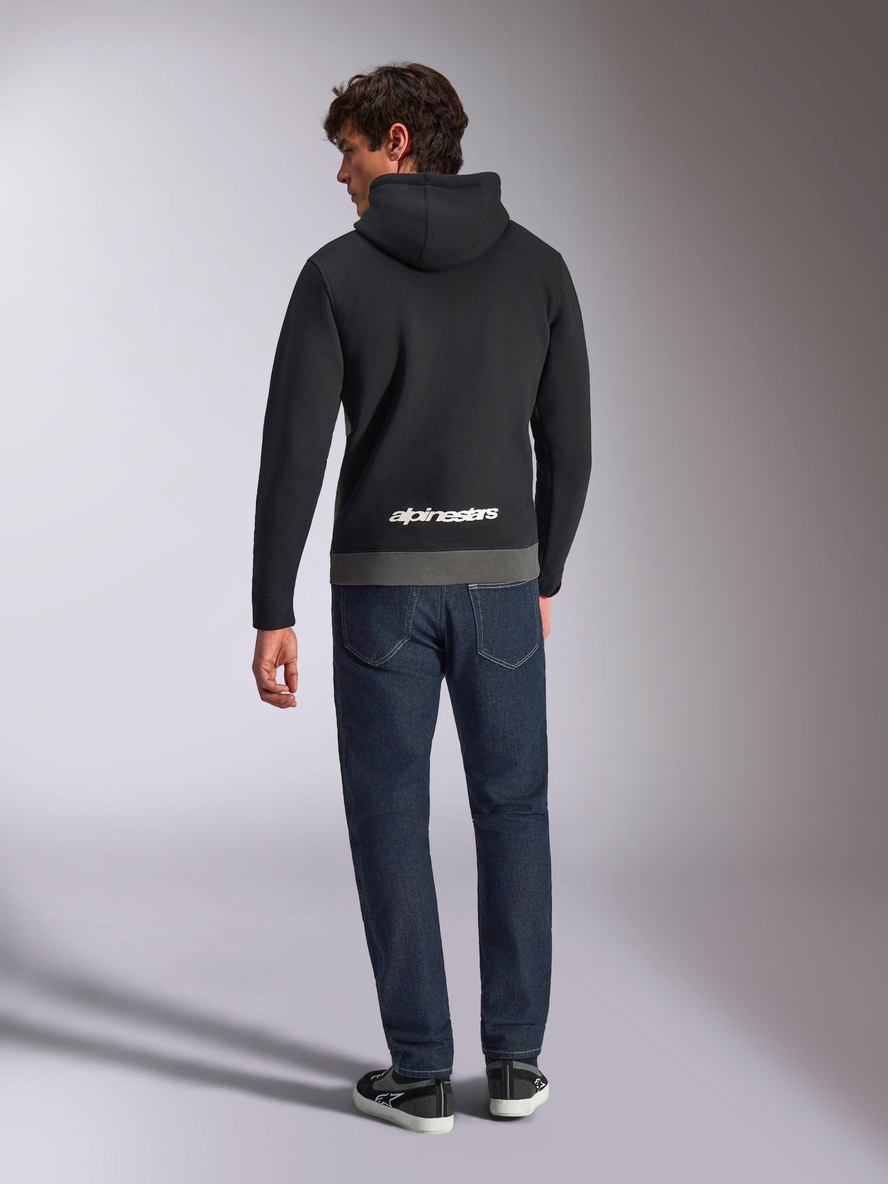Alpinestars Parallel Hoodie, Rückansicht eines Mannes in einem Fleece-Pullover mit Color-Blocking in Schwarz und Gunmetal, weißes Alpinestars-Wortlogo am unteren Rücken, kombiniert mit dunkelblauen Jeans, lässige Motorsport-inspirierte Streetwear