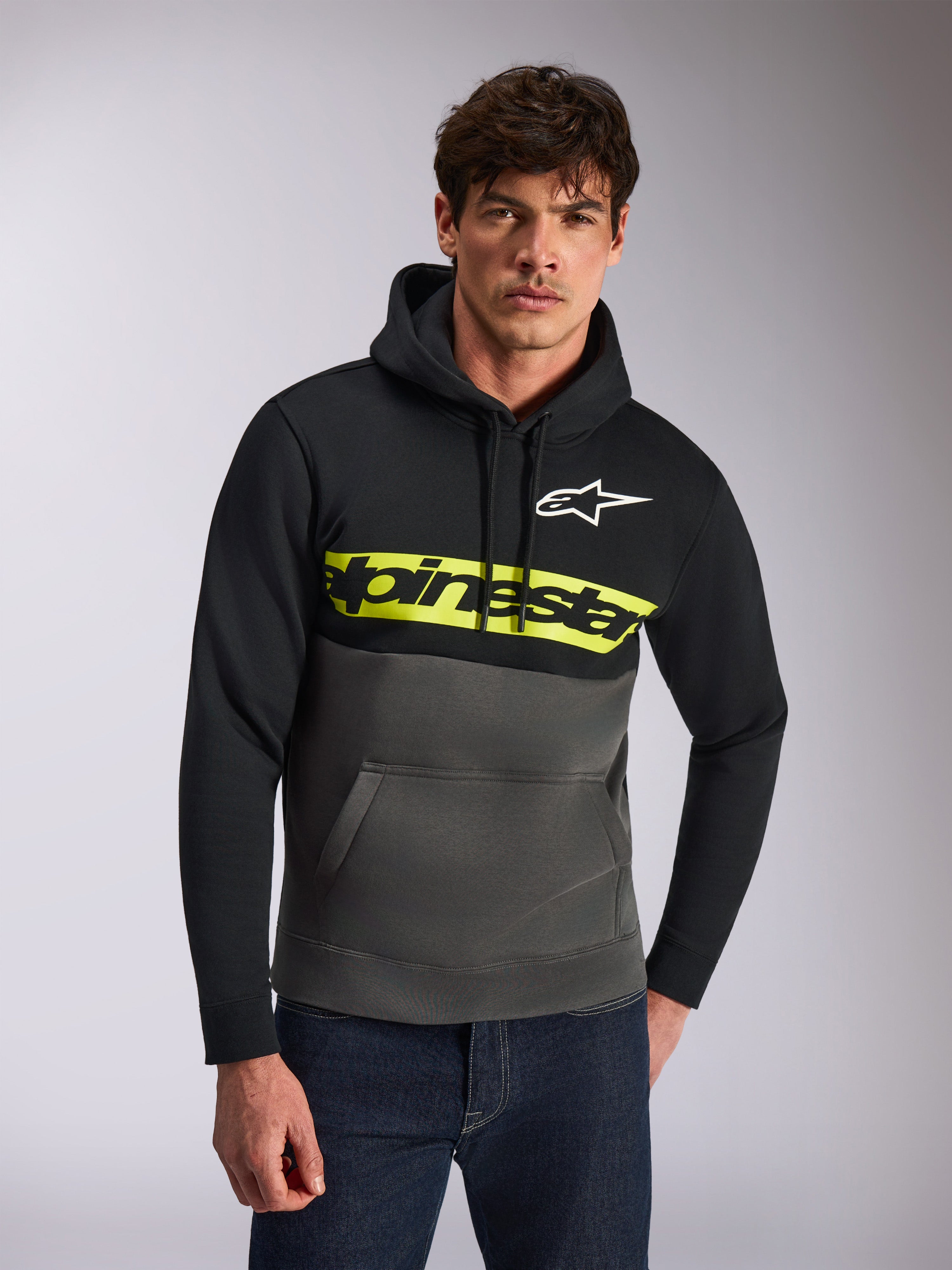 Alpinestars Parallel Hoodie, lässige Streetwear, Color-Blocking in Schwarz und Gunmetal-Grau, leuchtend gelber Bruststreifen mit Alpinestars-Wortlogo, weißes Astars-Logo, getragen von einem Model mit blauen Jeans, modernes vom Motorsport inspiriertes Design