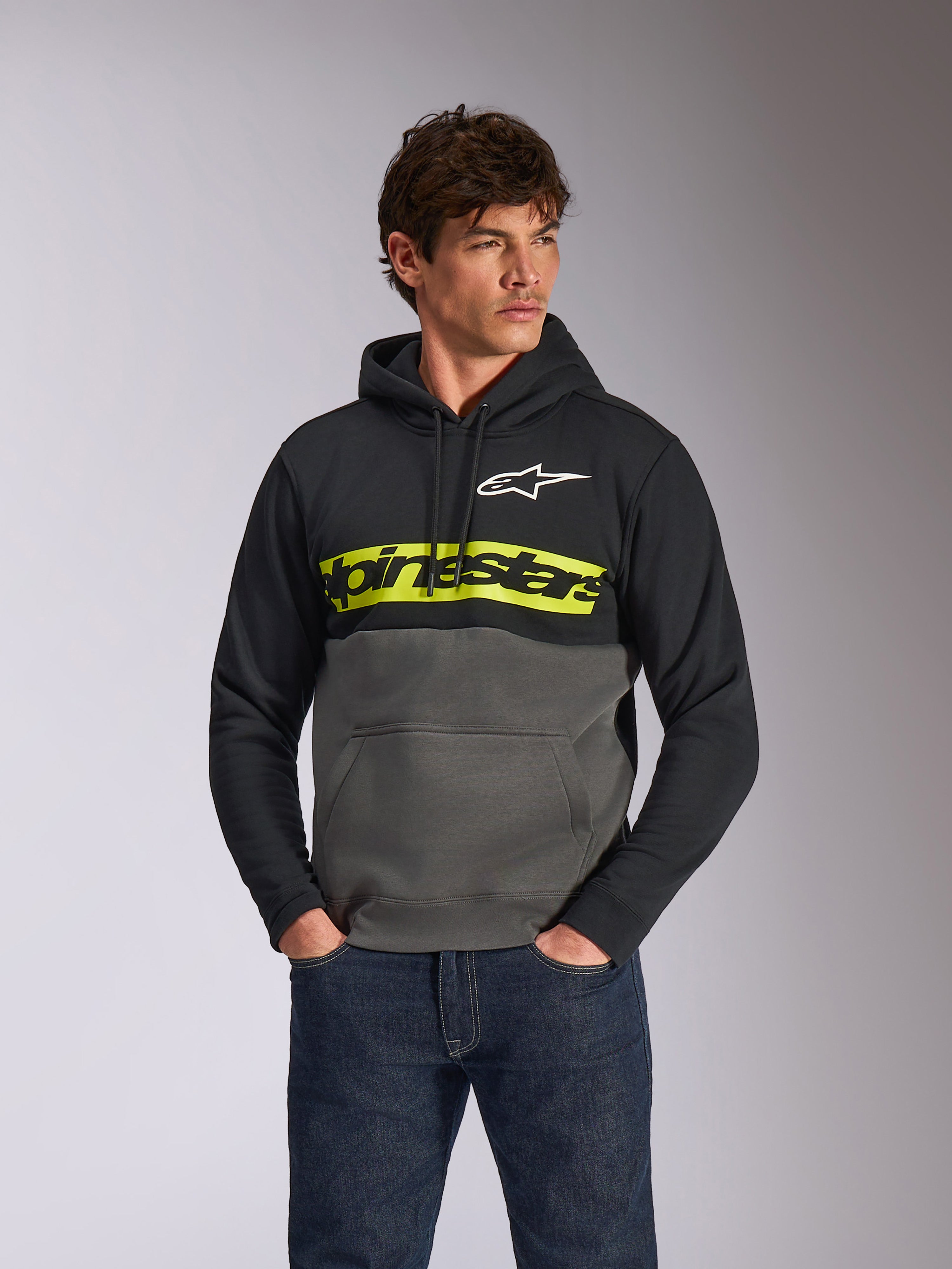 Alpinestars Parallel Hoodie, lässiger Fleece-Pullover im Color-Block-Design, Schwarz und Gunmetal mit einem leuchtend gelben Bruststreifen, Alpinestars Wortlogo und weißem Stern-Logo, getragen von einem männlichen Model mit dunkelblauen Jeans