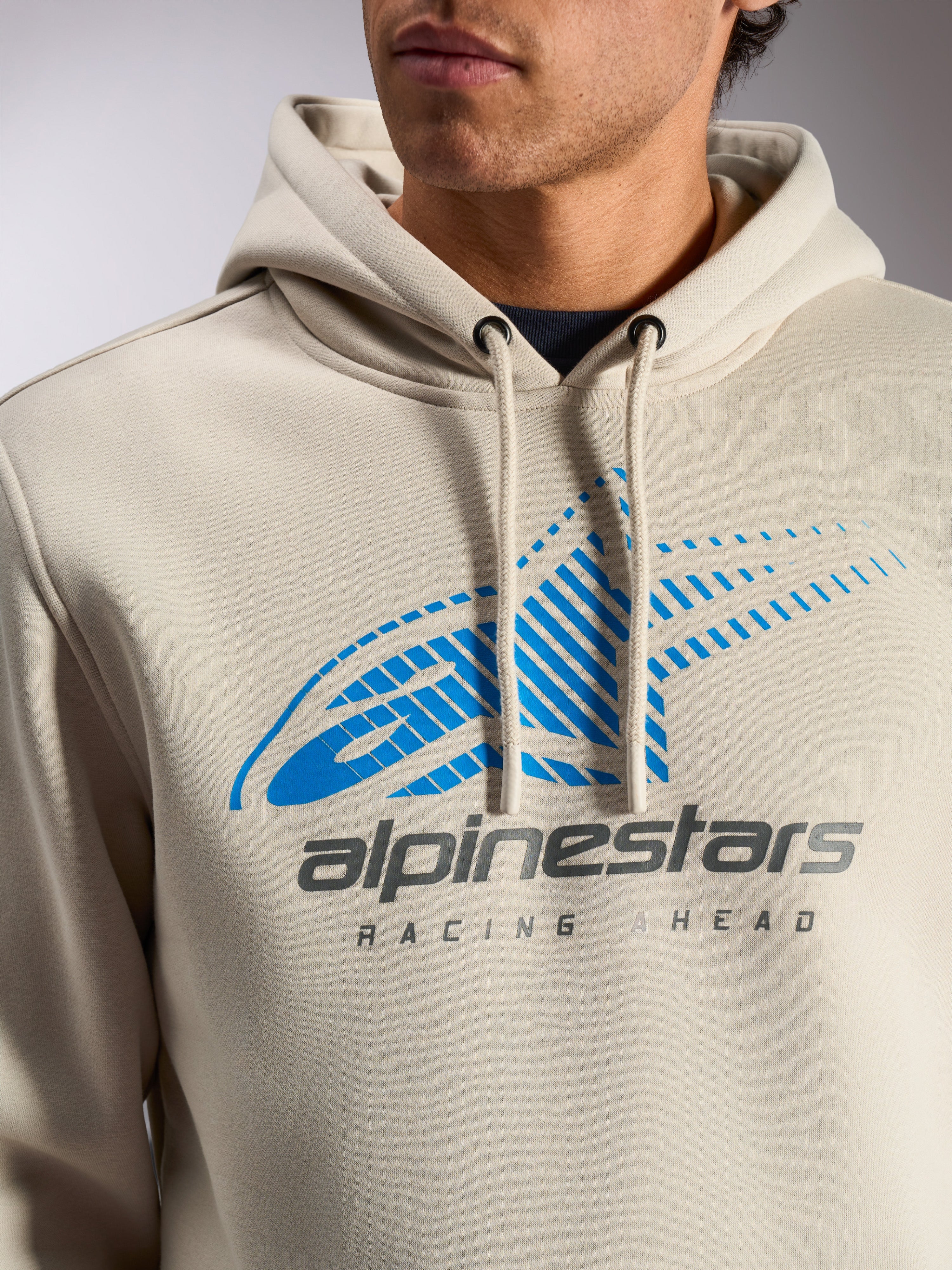 Alpinestars Pulse Hoodie, Off-White Casual-Streetwear-Pullover, Nahaufnahme der Brust mit blauem und grauem Astars Combo-Logo mit Speed-Line-Farbverlauf und „Racing Ahead“-Schriftzug, Kapuze mit Kordelzug und Kordelenden mit Tauchspitze.