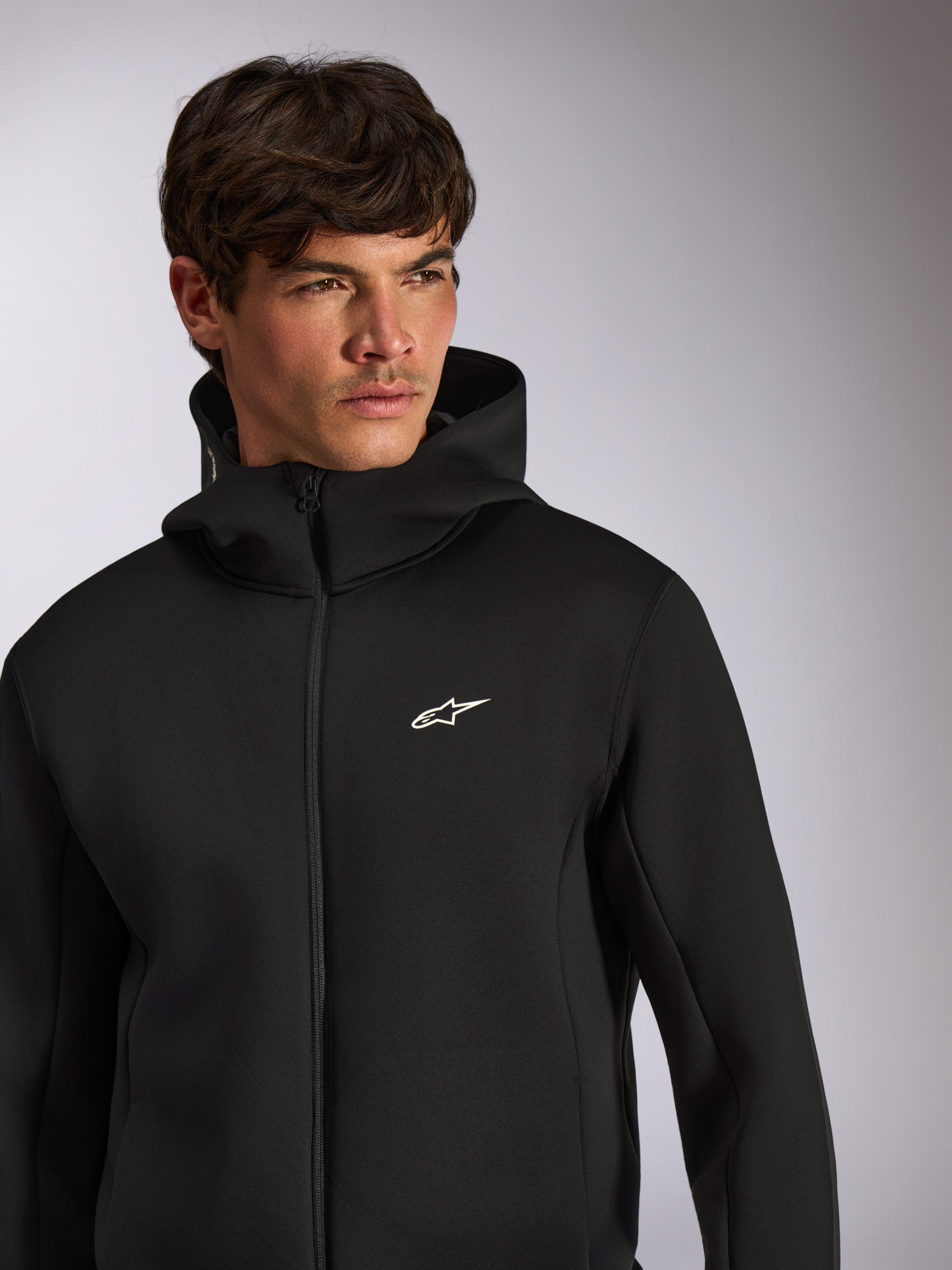 Alpinestars Cypher Hoodie, lässige Sportbekleidung, schwarzer Hoodie mit Frontreißverschluss, weißes Astars-Logo auf der Brust, Kapuzendesign mit hohem Kragen, getragen von einem männlichen Model