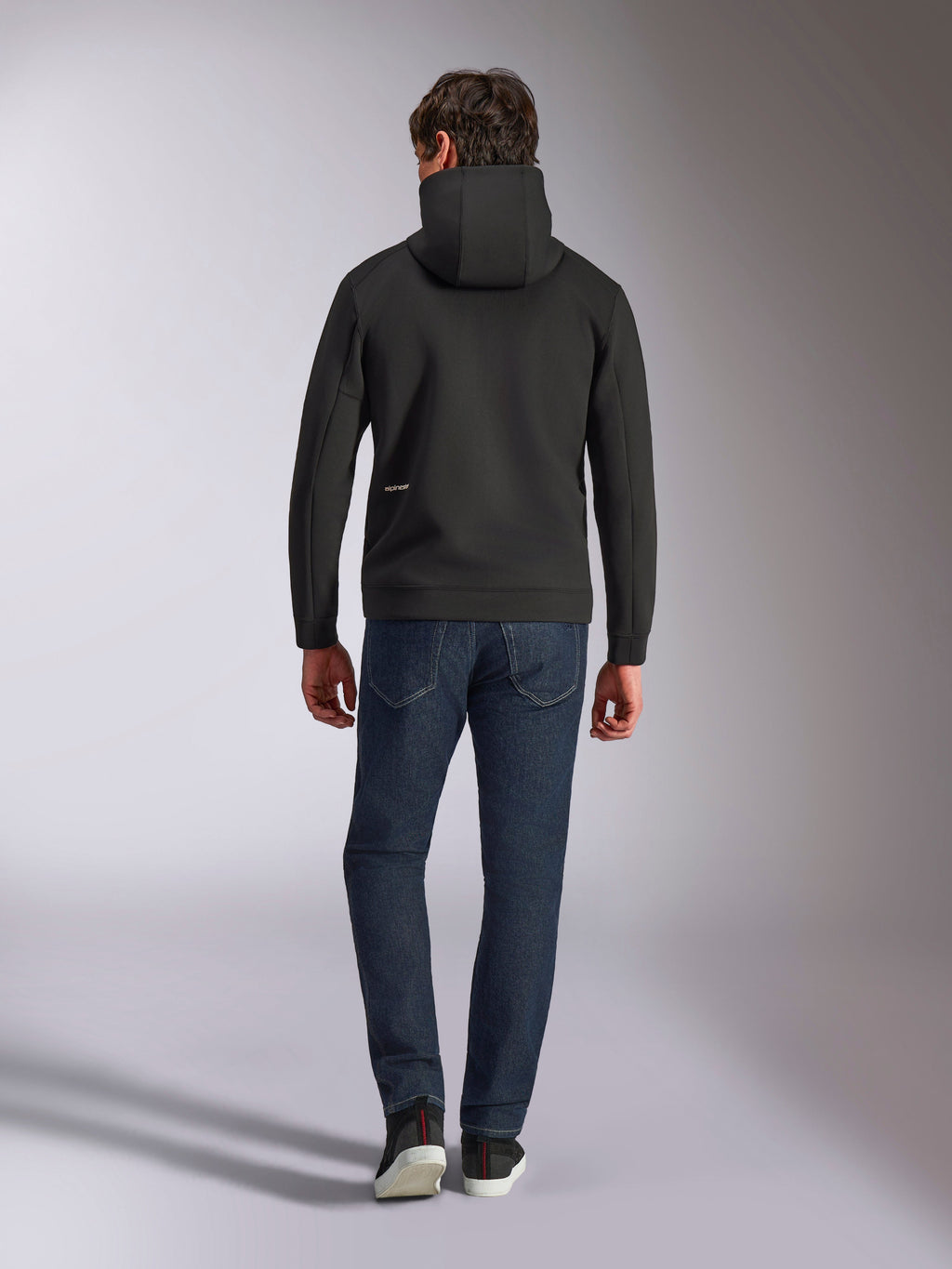 Alpinestars Cypher Hoodie, schwarzer Doppelvlies-Kapuzenpullover, Rückansicht mit kleinem weißen Alpinestars-Logo auf der unteren linken Hüfte, Kapuze mit hohem Kragen, kombiniert mit dunkelblauer Jeans, lässige vom Motorsport inspirierte Streetwear