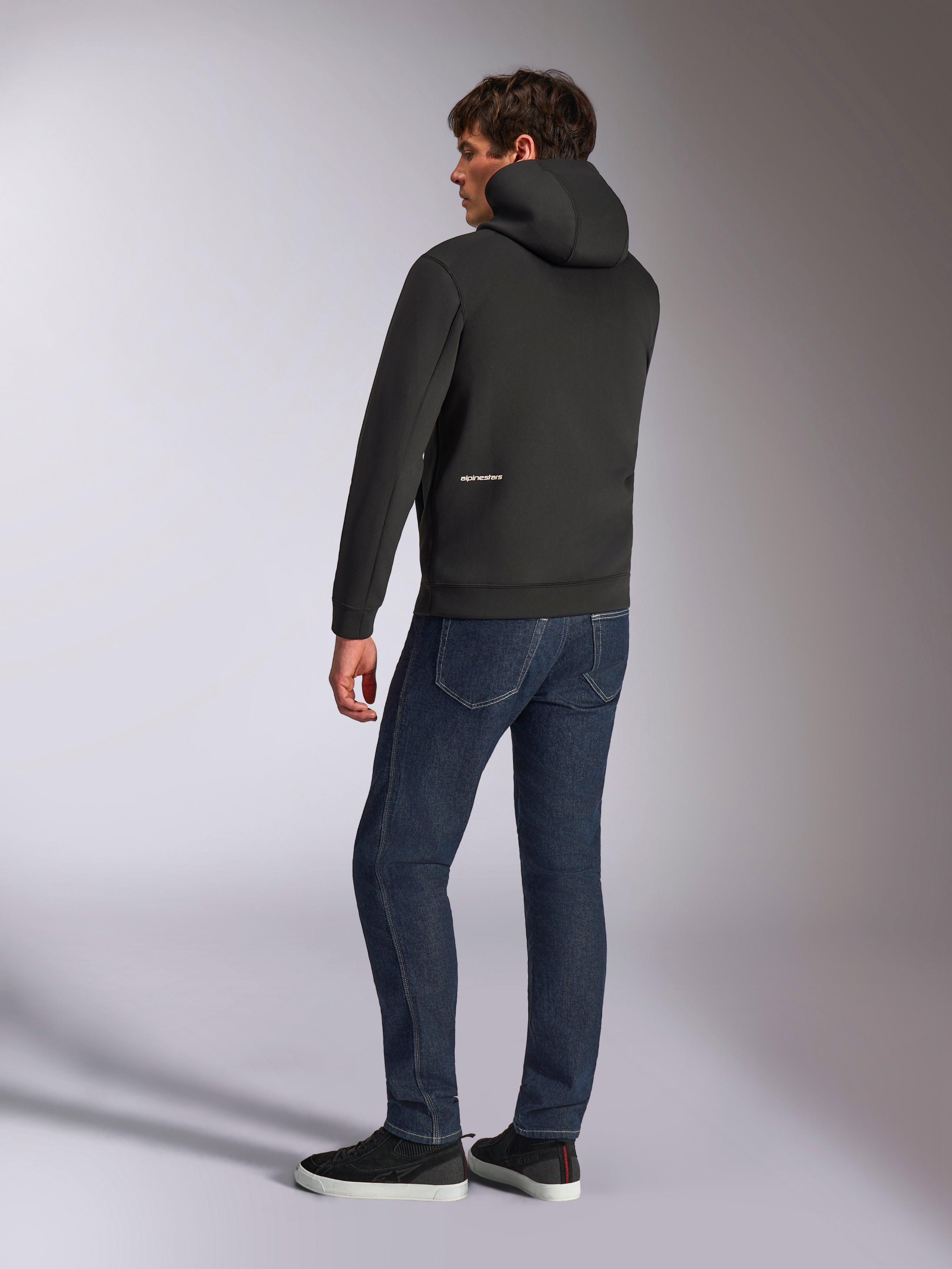 Alpinestars Cypher Hoodie, schwarzer lässiger Zip-Hoodie, Rückansicht mit hochgeschlossener Kapuze und weißem Alpinestars-Logo am unteren Rücken, kombiniert mit dunkelblauer Jeans und schwarzen Sneakern, modernes, vom Motorsport inspiriertes Design
