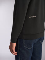 Alpinestars Cypher Hoodie, Nahaufnahme des unteren Rückens und Ärmels, schwarzes Doppelvlies mit weißem Alpinestars-Wortlogo, getragen mit blauen Jeans, lässige Motorsport-Streetwear