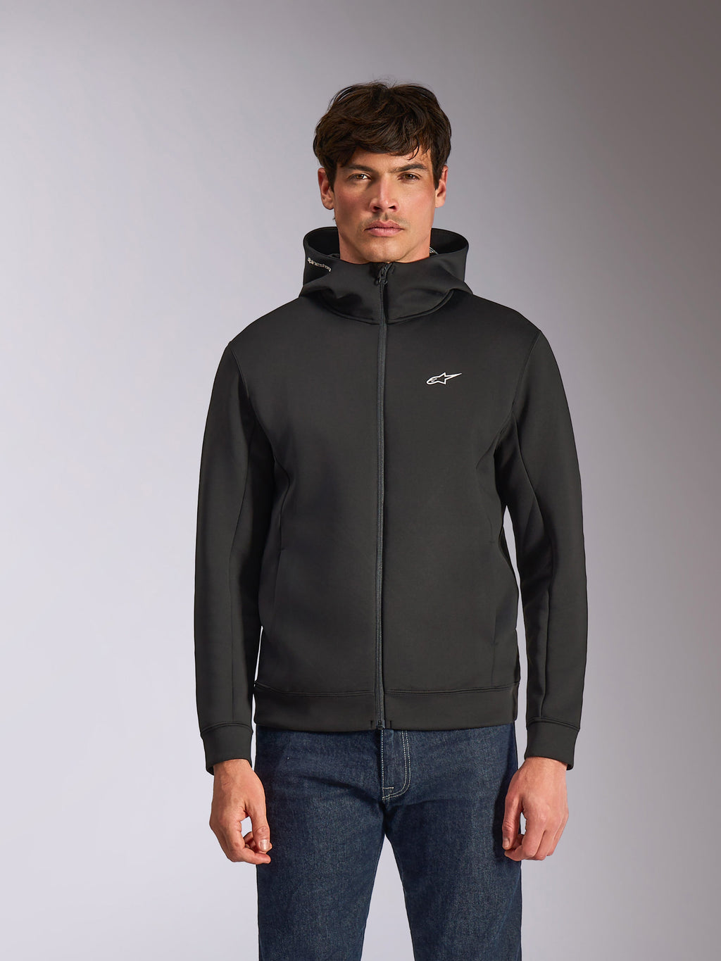 Alpinestars Cypher Hoodie, schwarzer Fleece-Hoodie mit Frontreißverschluss, Kapuze mit hohem Kragen und weißem Branding, kleines weißes Astars-Logo auf der Brust, getragen von einem Model mit blauen Jeans, lässige vom Motorsport inspirierte Streetwear.