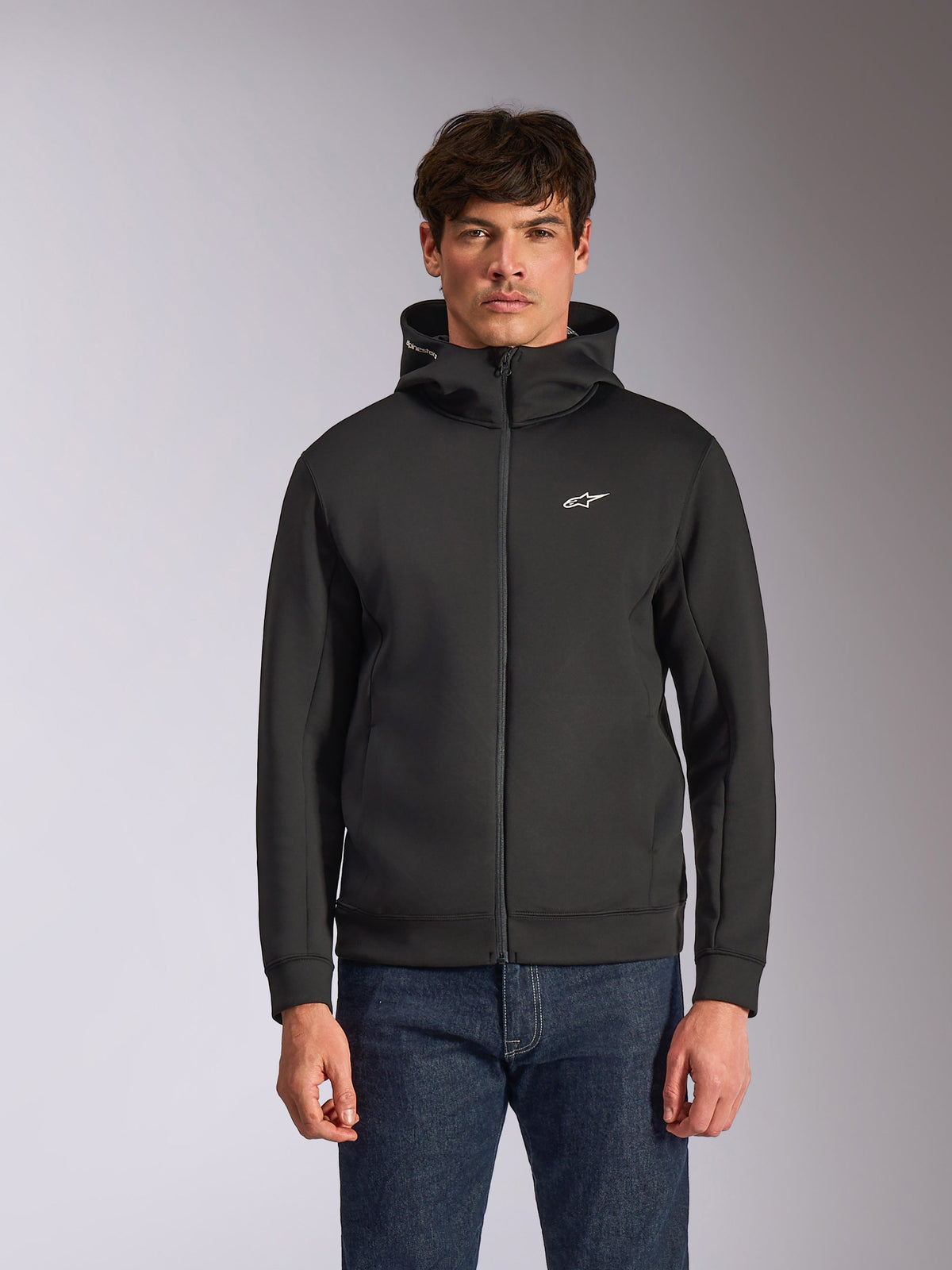 Alpinestars Cypher Hoodie, schwarzer Fleece-Hoodie mit Frontreißverschluss, Kapuze mit hohem Kragen und weißem Branding, kleines weißes Astars-Logo auf der Brust, getragen von einem Model mit blauen Jeans, lässige vom Motorsport inspirierte Streetwear.