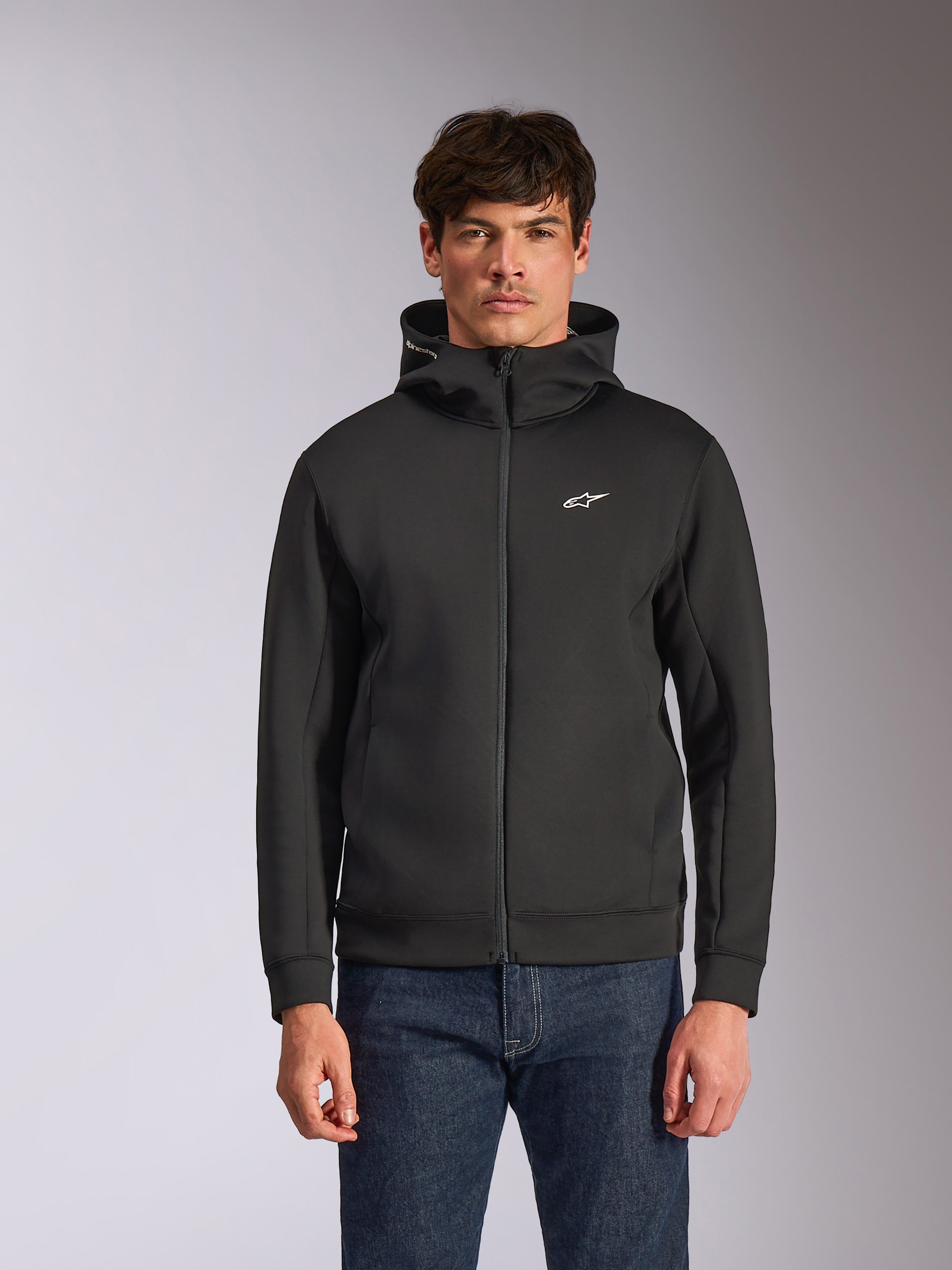 Alpinestars Cypher Hoodie, schwarzer Fleece-Hoodie mit Frontreißverschluss, Kapuze mit hohem Kragen und weißem Branding, kleines weißes Astars-Logo auf der Brust, getragen von einem Model mit blauen Jeans, lässige vom Motorsport inspirierte Streetwear.