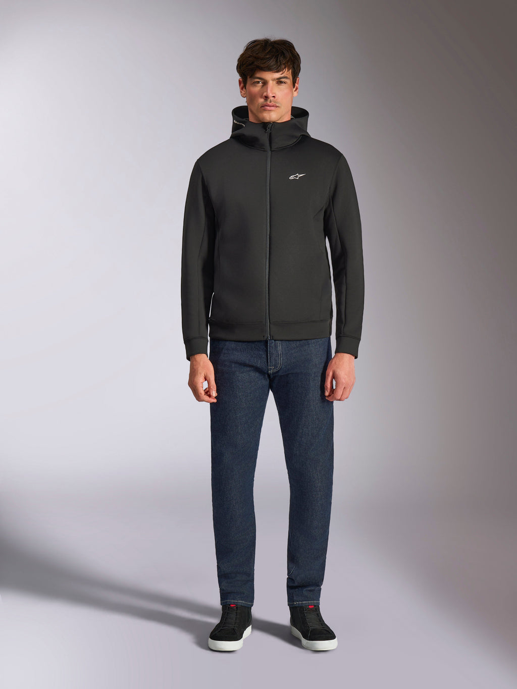 Modell trägt den Alpinestars Cypher Hoodie, schwarzer Fleece-Hoodie mit Front-Reißverschluss, hochgeschlossene Kapuze mit kleinem weißen Astars-Logo auf der Brust, kombiniert mit dunkler Denim-Jeans und schwarzen Sneakern, lässige Motorsport-inspirierte Streetwear